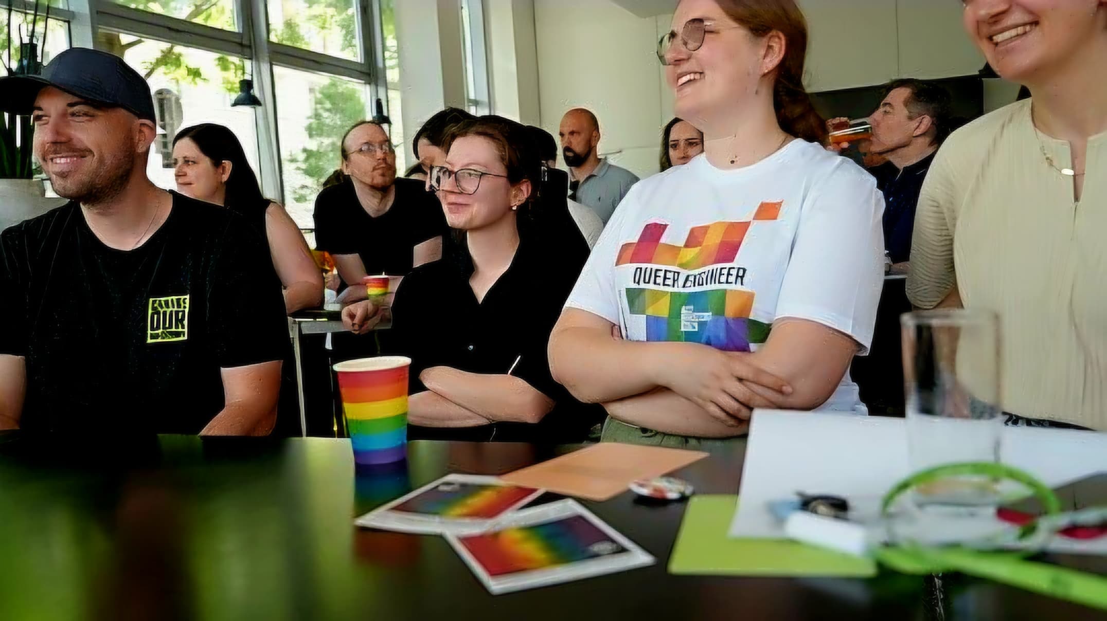 Eine Gruppe von Menschen sitzt in einem hellen Raum und lächelt. Eine Person im Vordergrund trägt ein „Queer Engineer“-T-Shirt mit einer Regenbogenfahne. Auf dem Tisch stehen Tassen und Karten mit Regenbogenmotiven, was auf eine LGBTQ+-inklusive Veranstaltung hindeutet.