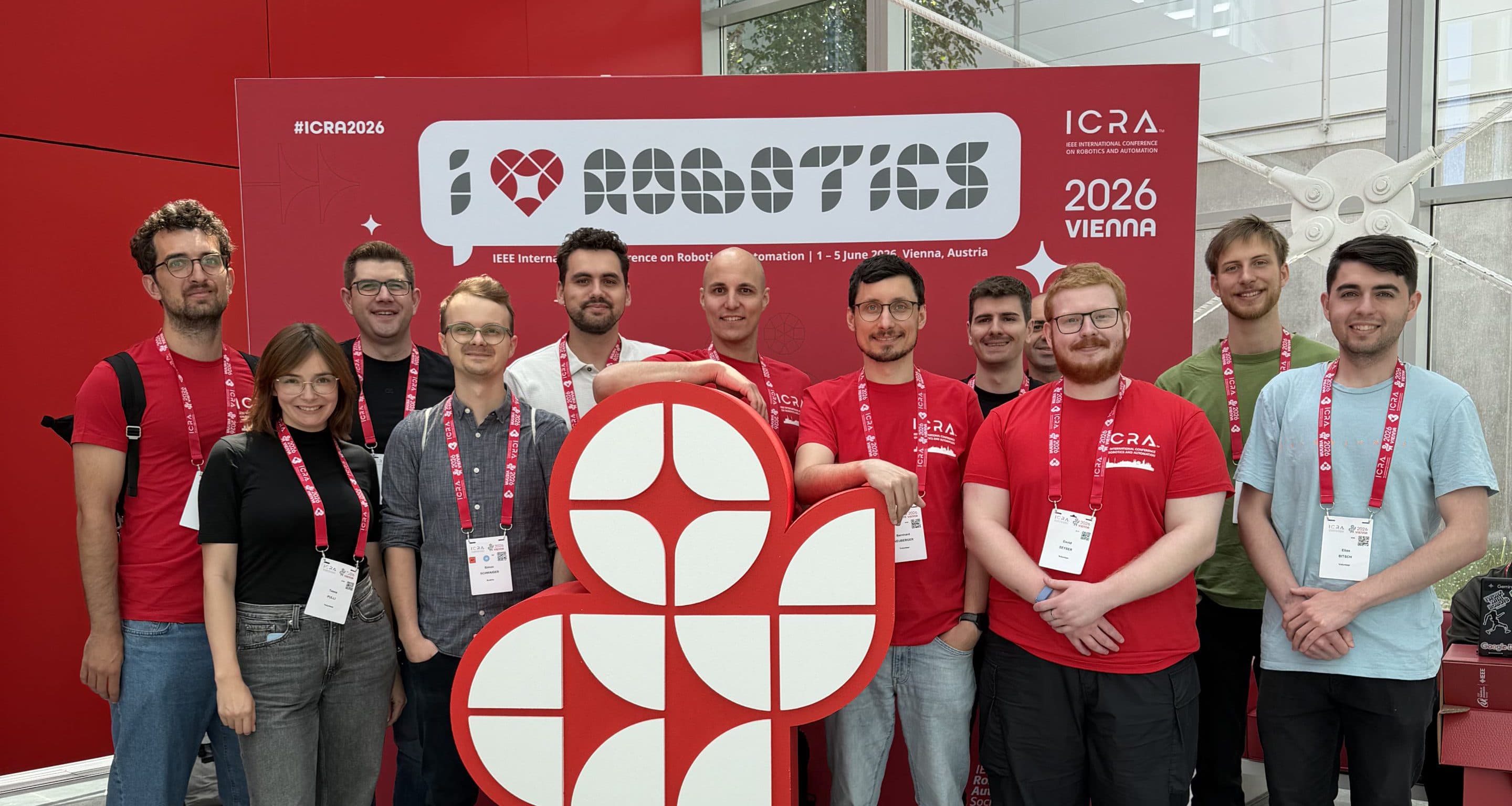 Eine Gruppe von Menschen mit Konferenzausweisen und roten T-Shirts steht gemeinsam in einem Raum vor einem großen „I ♥ Robotics“-Schild auf der ICRA 2026 in Wien.