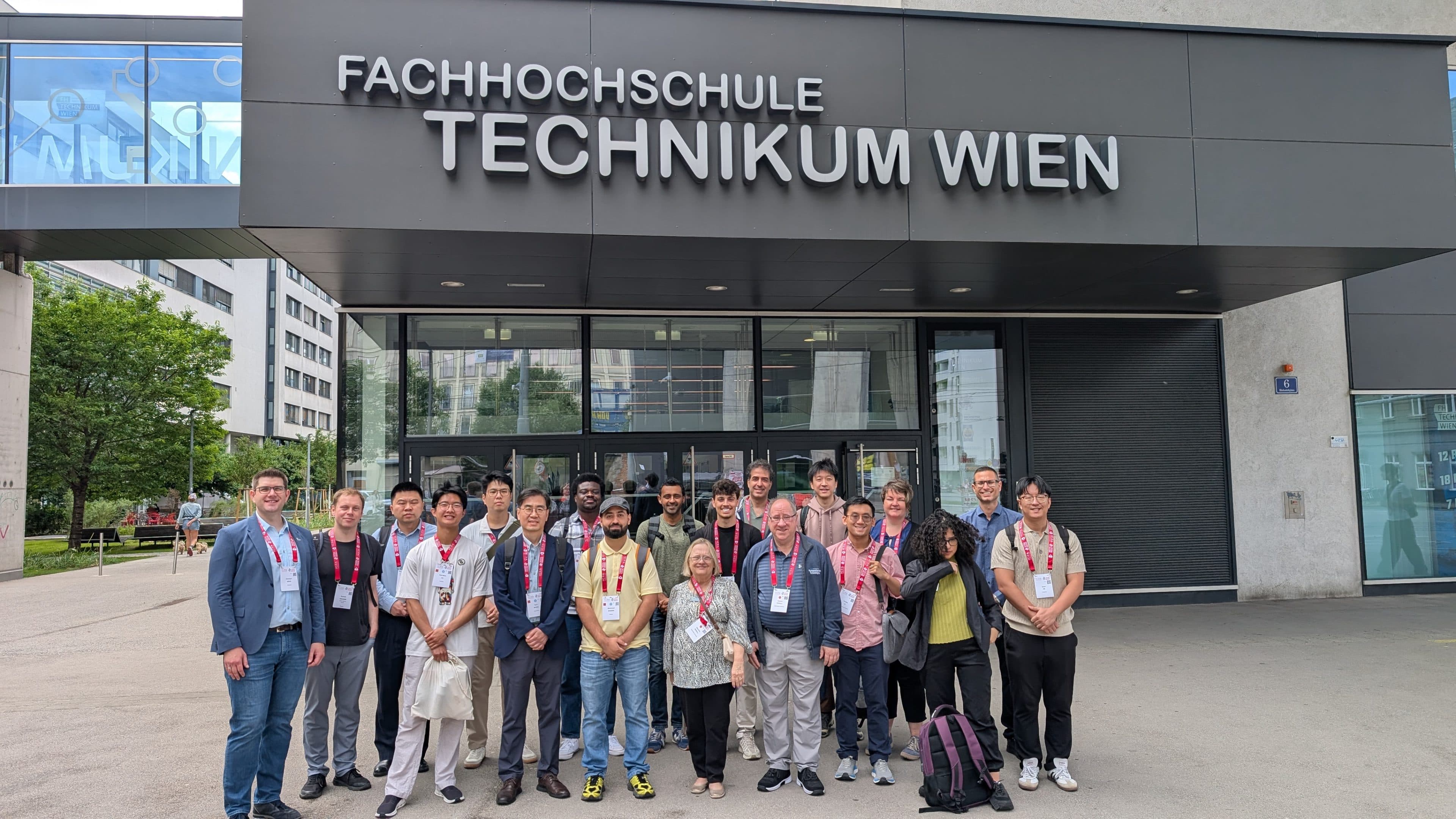 Eine Gruppe von Menschen steht lächelnd vor dem Eingang der Fachhochschule Technikum Wien, einem modernen Hochschulgebäude mit großen Glasfenstern, über dessen Eingang der Name der Hochschule prangt.