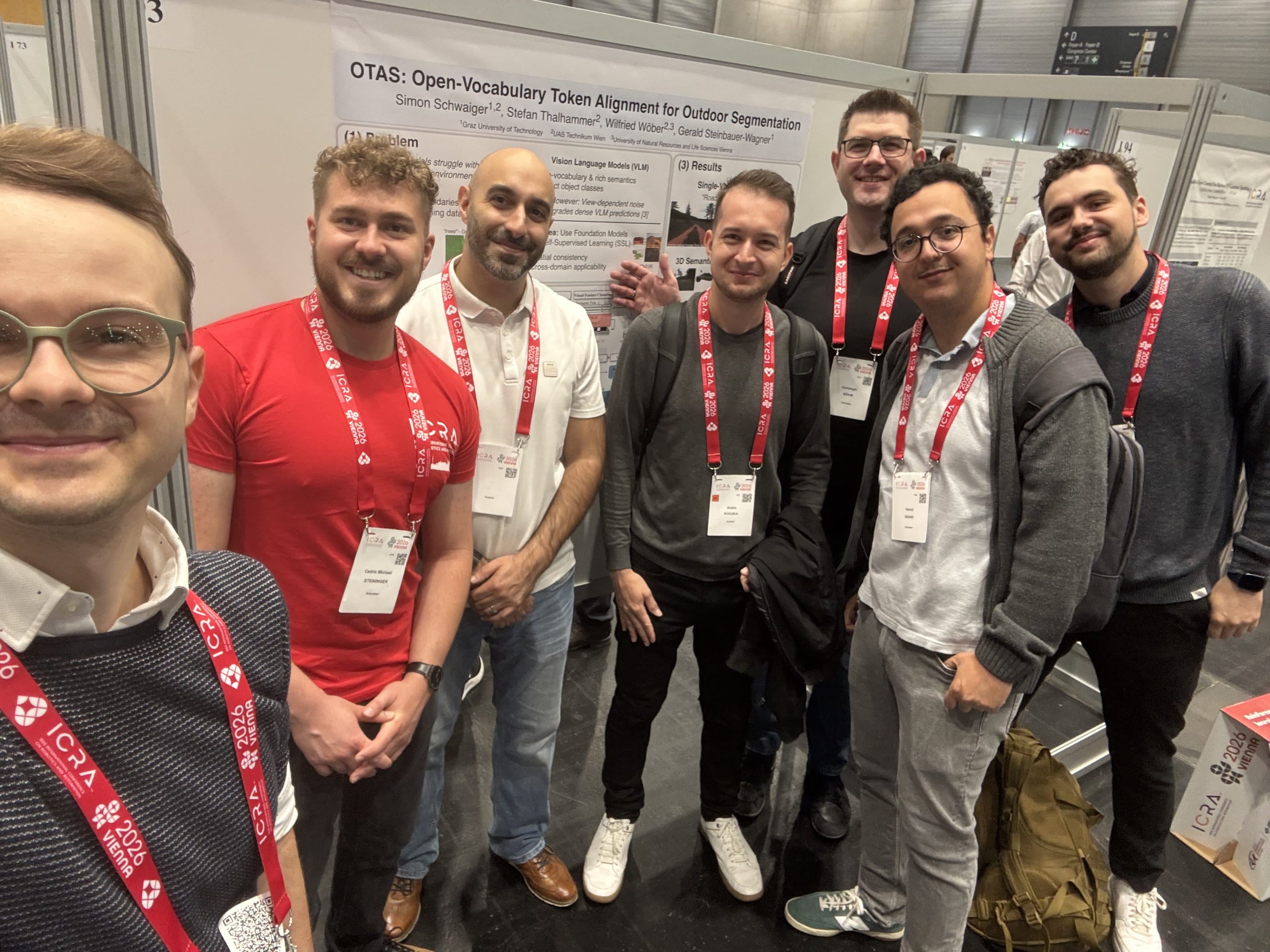 Sieben Personen stehen lächelnd für ein Gruppen-Selfie vor einem wissenschaftlichen Poster mit dem Titel „OTAS: Open-Vocabulary Token Alignment for Outdoor Segmentation“ – offenbar im Rahmen einer Konferenz oder einer wissenschaftlichen Veranstaltung.