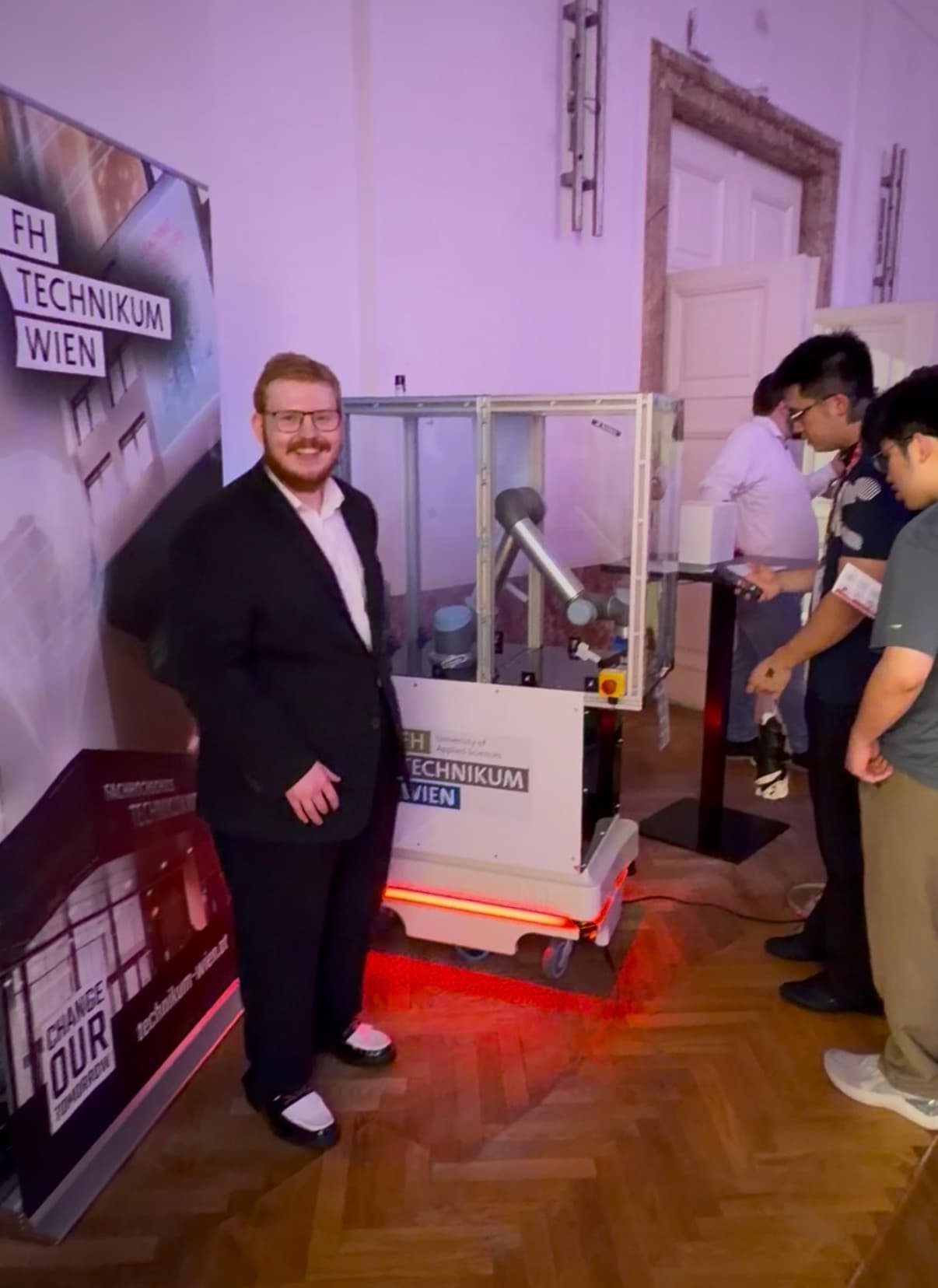 Bei einer Veranstaltung des FH Technikum Wien steht ein Mann im Anzug lächelnd neben einem Roboterarm-Exponat, während andere Besucher in einem hell beleuchteten Raum mit Holzboden mit der Ausstellung interagieren.