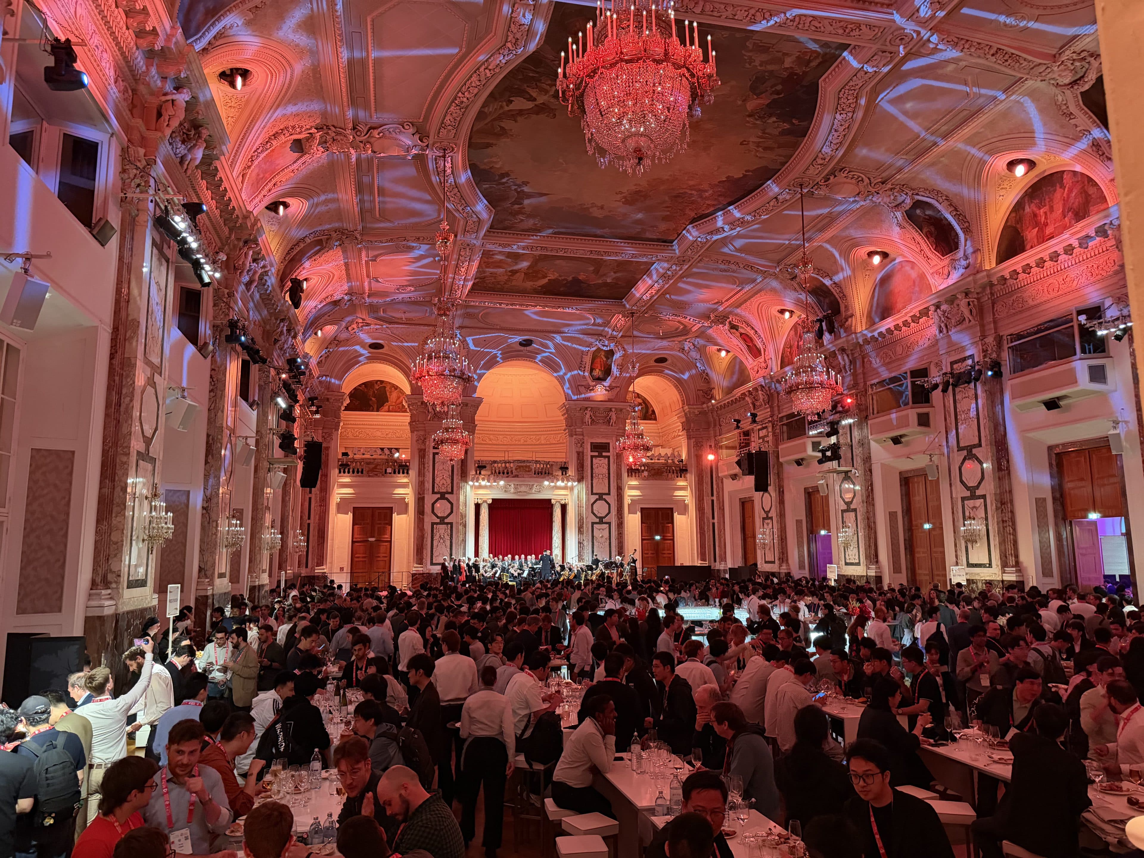 Ein großer, eleganter Ballsaal, in dem die Gäste an Tischen sitzen, an der Decke hängen kunstvolle Kronleuchter, und bunte Lichter setzen die dekorative Architektur in Szene. Am anderen Ende des Raums ist eine Bühne zu sehen.