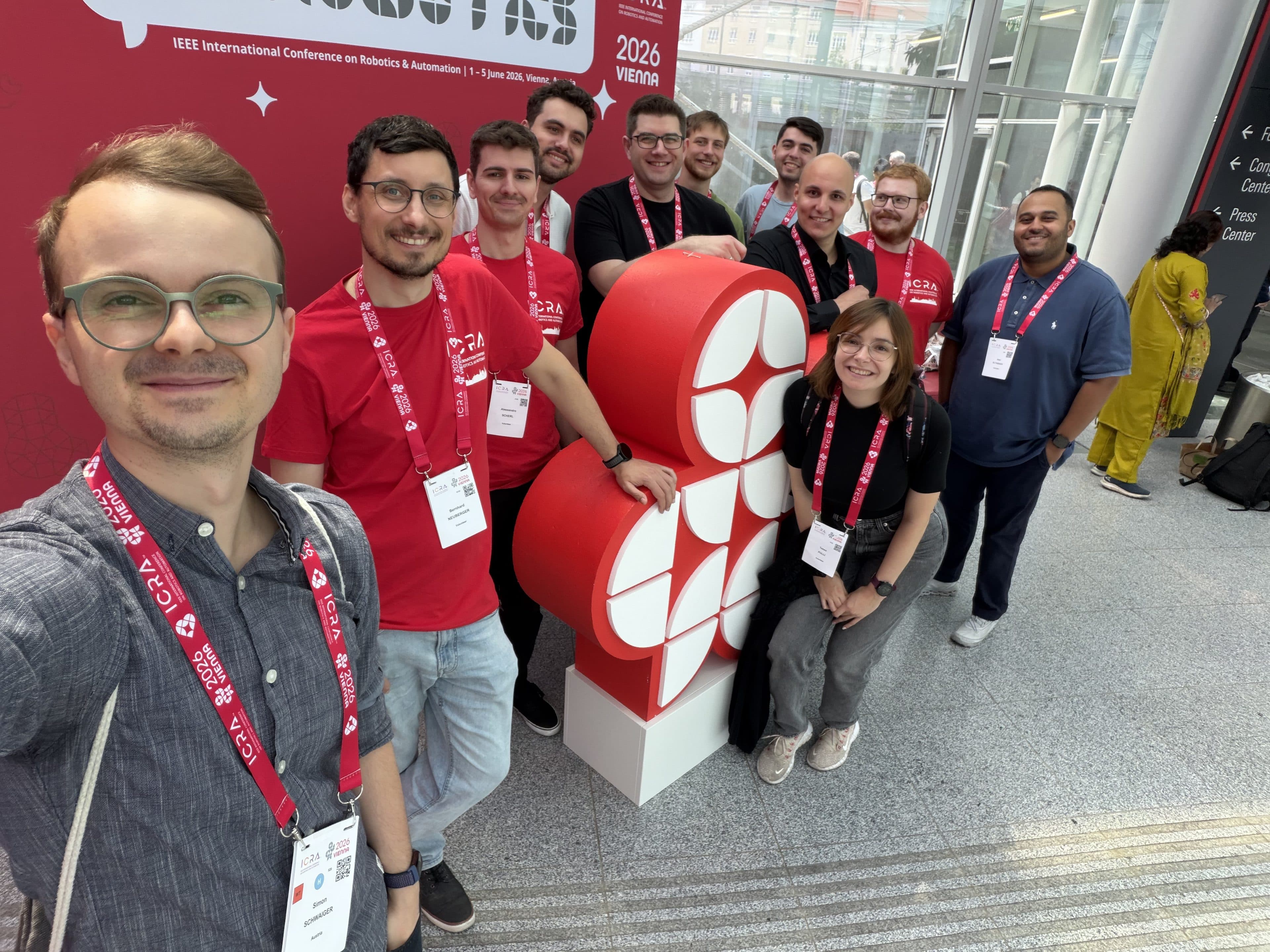 Eine Gruppe von Menschen mit Konferenzausweisen posiert lächelnd vor einer roten, herzförmigen Skulptur auf der IEEE International Conference on Robotics & Automation 2024, die in einer modernen Halle stattfindet.