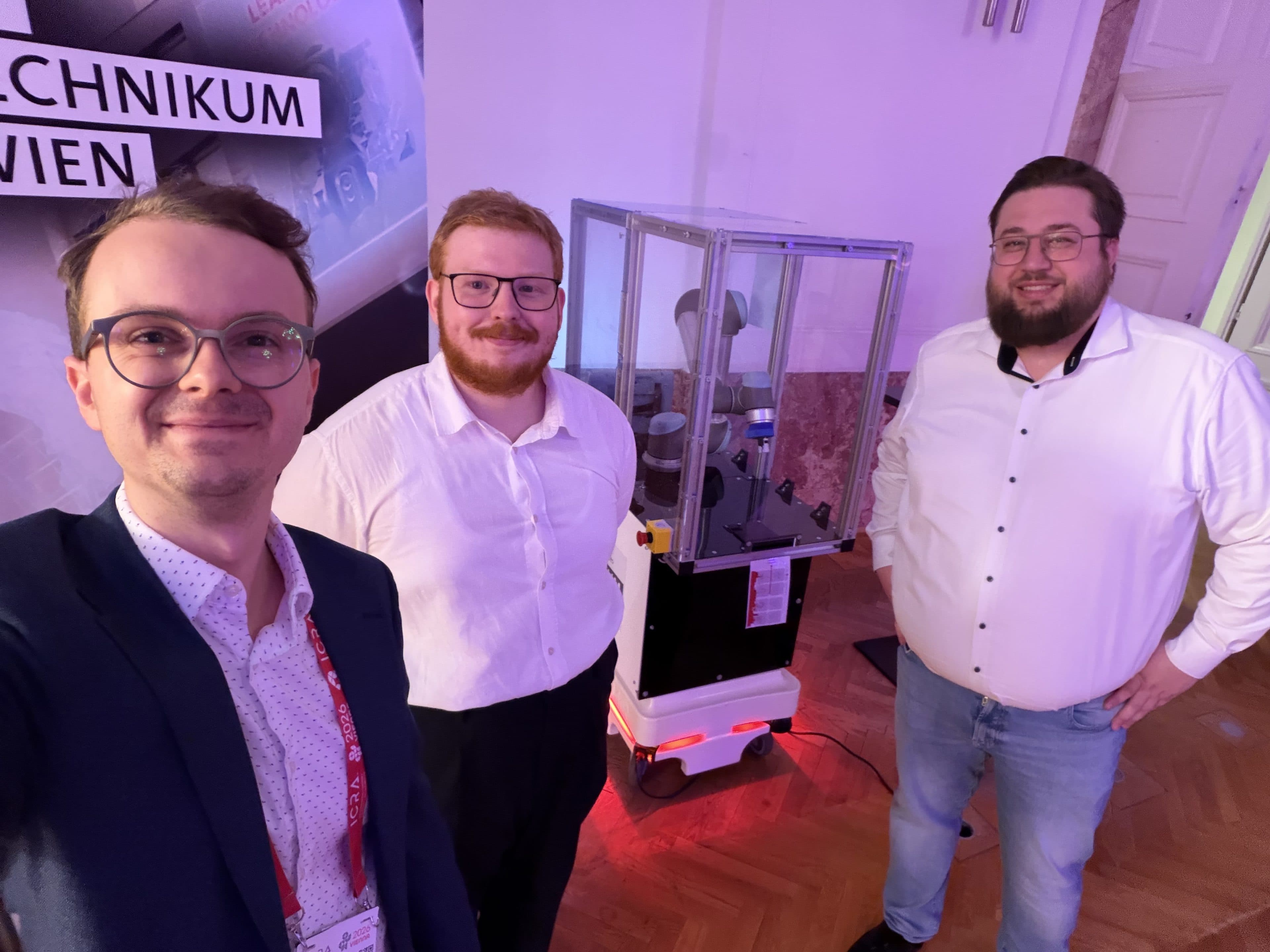 Drei Männer in Geschäftskleidung lächeln für ein Selfie in einem Innenraum. Hinter ihnen sind ein Robotergerät und ein Banner mit teilweise sichtbarem Text zu sehen, was auf eine Veranstaltung aus dem Technologie- oder Wissenschaftsbereich hindeutet.