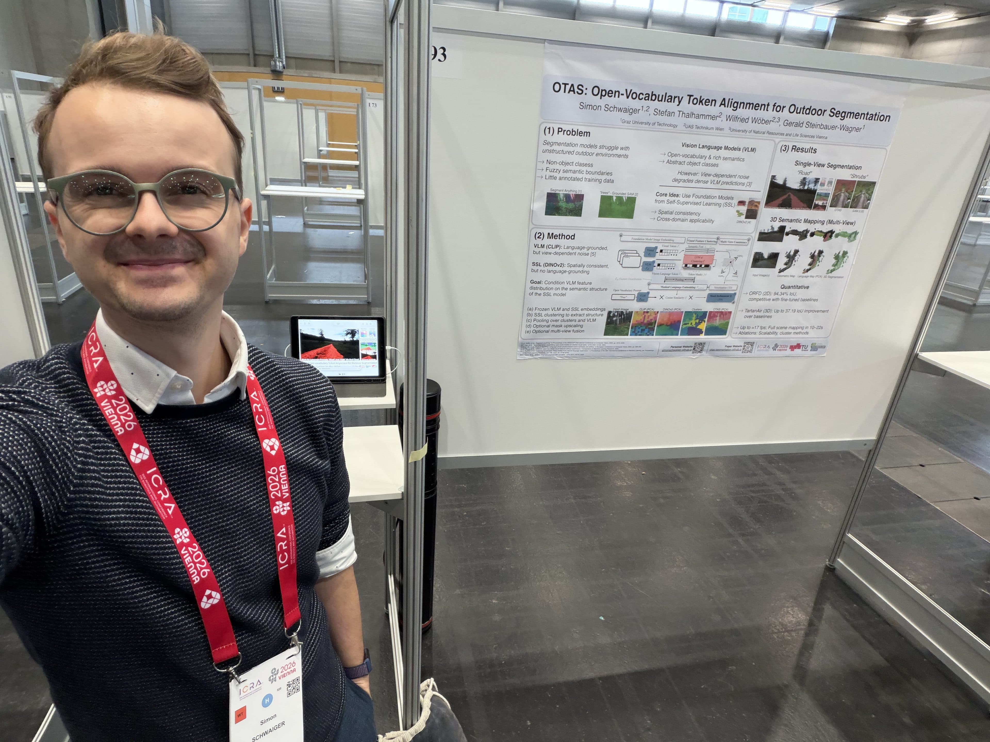 Eine lächelnde Person mit Brille und einem roten Konferenz-Lanyard macht vor einem Forschungsplakat mit dem Titel „OTAS: Open-Vocabulary Token Alignment for Outdoor Segmentation“ in einer Konferenz- oder Ausstellungshalle ein Selfie.