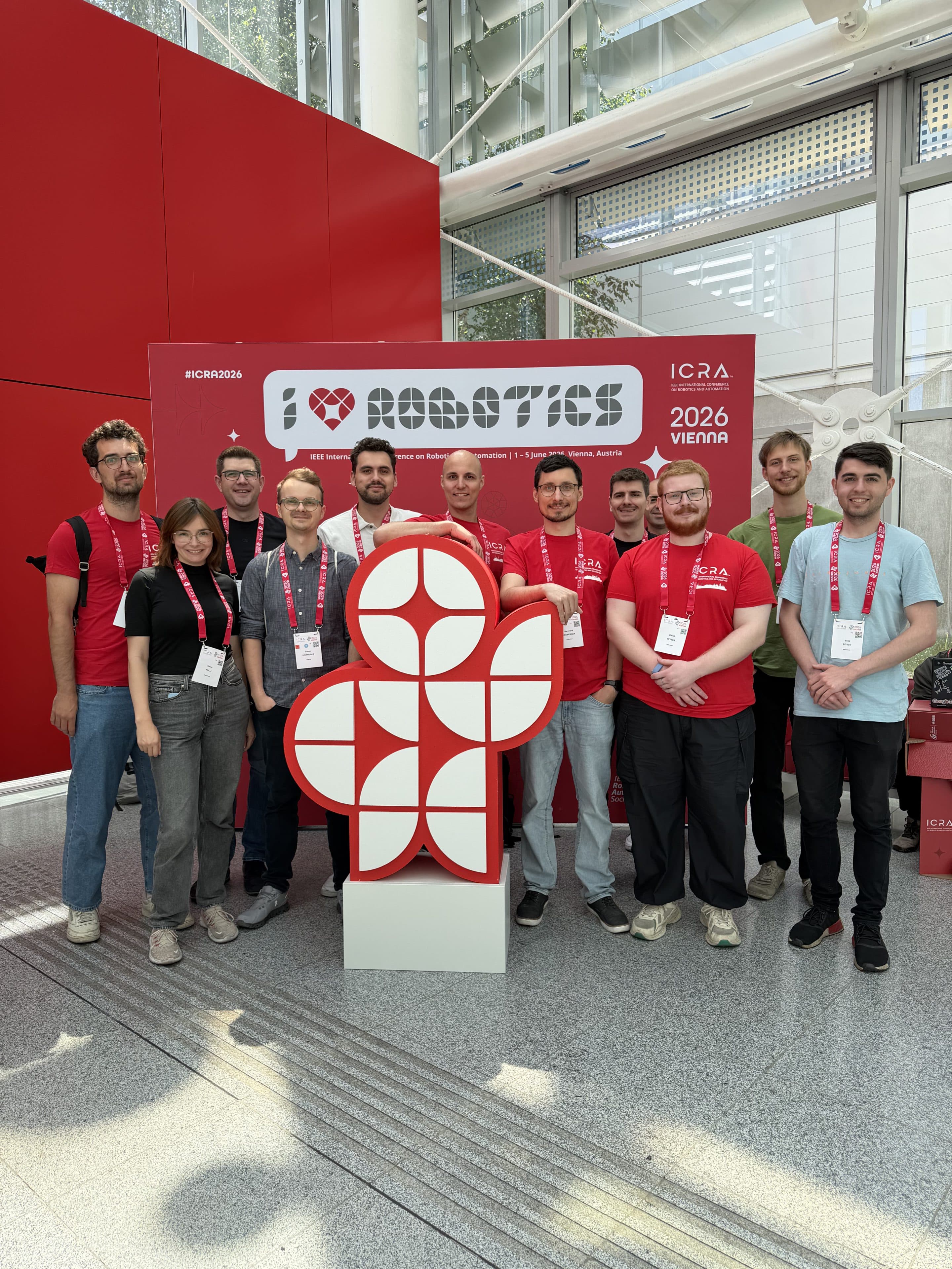 Bei einer Veranstaltung in einer Halle steht eine Gruppe von elf Personen lächelnd um ein Schild mit einem geometrischen Logo und der Aufschrift „I ♥ ROBOTICS“ herum, im Hintergrund sind Banner für die ICRA 2026 in Wien zu sehen.