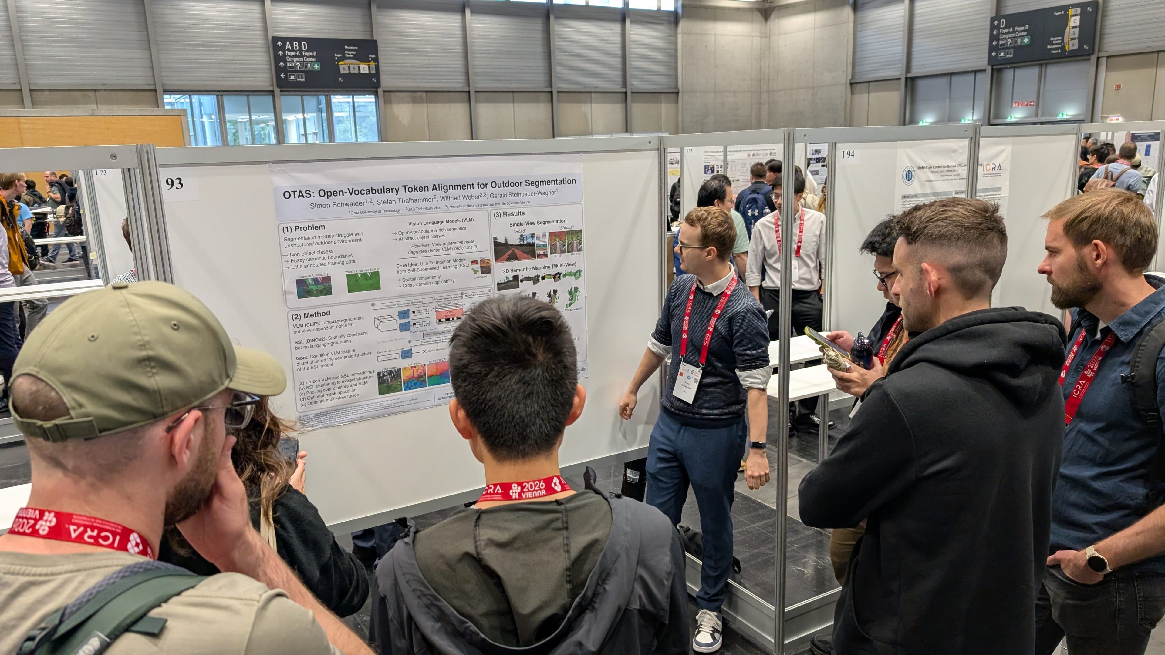 Auf einer Konferenz versammelt sich eine Gruppe von Menschen um einen Referenten, der ein wissenschaftliches Poster mit dem Titel „OTAS: Open-Vocabulary Token Alignment for Outdoor Segmentation“ erläutert. Der Referent deutet auf das Poster, während die Teilnehmer zuhören.
