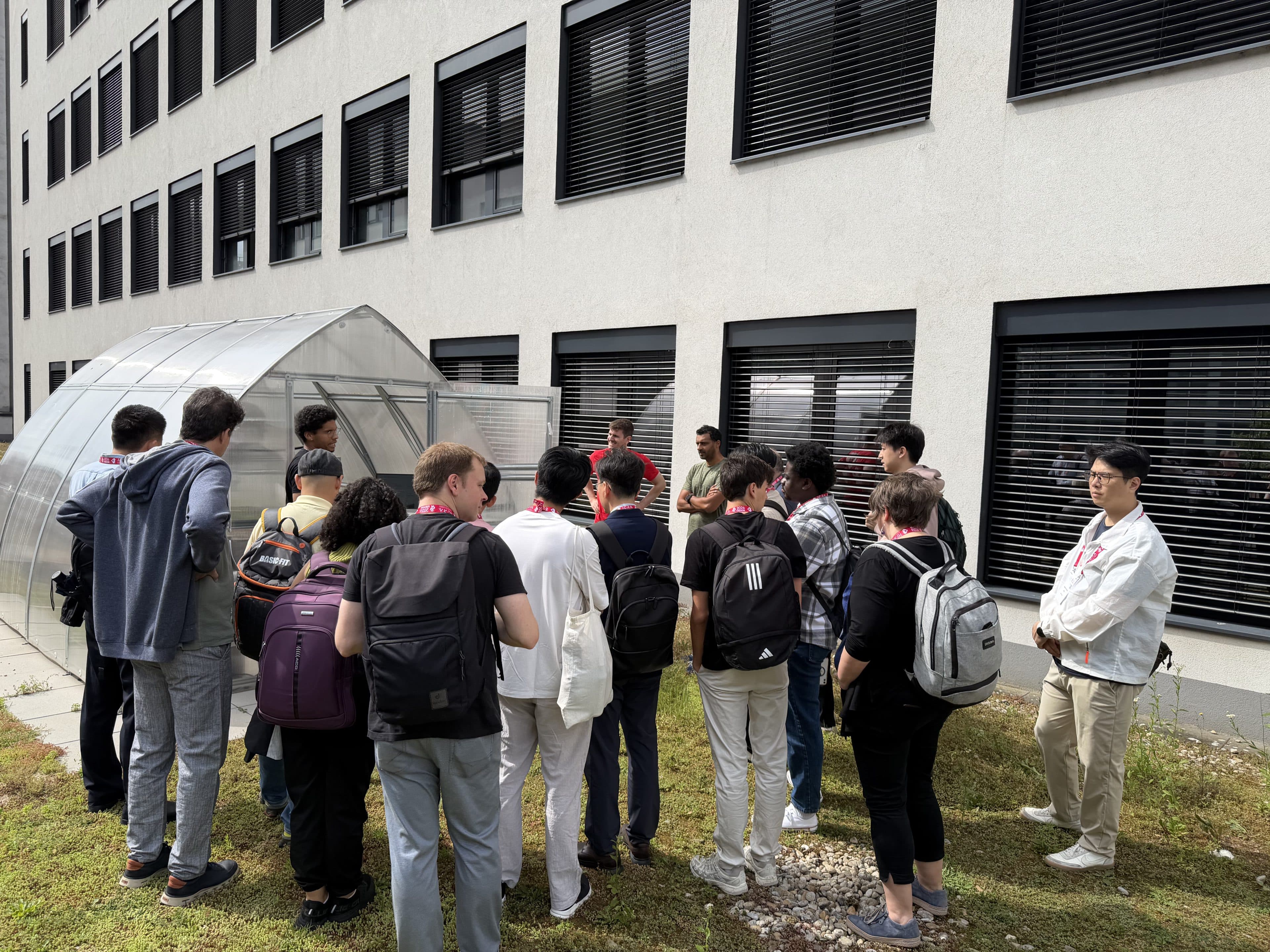 Eine Gruppe von Menschen mit Rucksäcken steht vor einem Gebäude mit großen Fenstern und schwarzen Fensterläden und hört jemandem zu, der in der Nähe eines kleinen Gewächshauses auf einer Rasenfläche spricht.