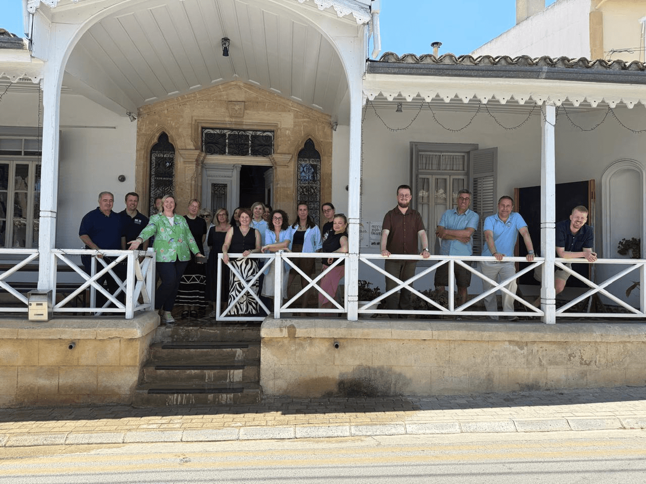 Eine Gruppe von Menschen steht an einem sonnigen Tag auf der Veranda eines historisch anmutenden Gebäudes mit weißen Geländern und Säulen und lächelt.