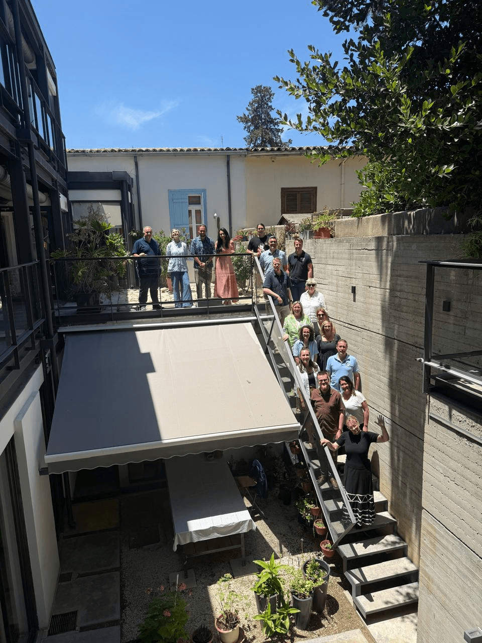 Eine Gruppe von Menschen steht gemeinsam auf einer Treppe und einem Balkon in einem sonnendurchfluteten Innenhof und posiert für ein Foto. Einige winken in die Kamera. Im Innenhof stehen Topfpflanzen und ein Tisch, der von einer Markise überdacht ist.