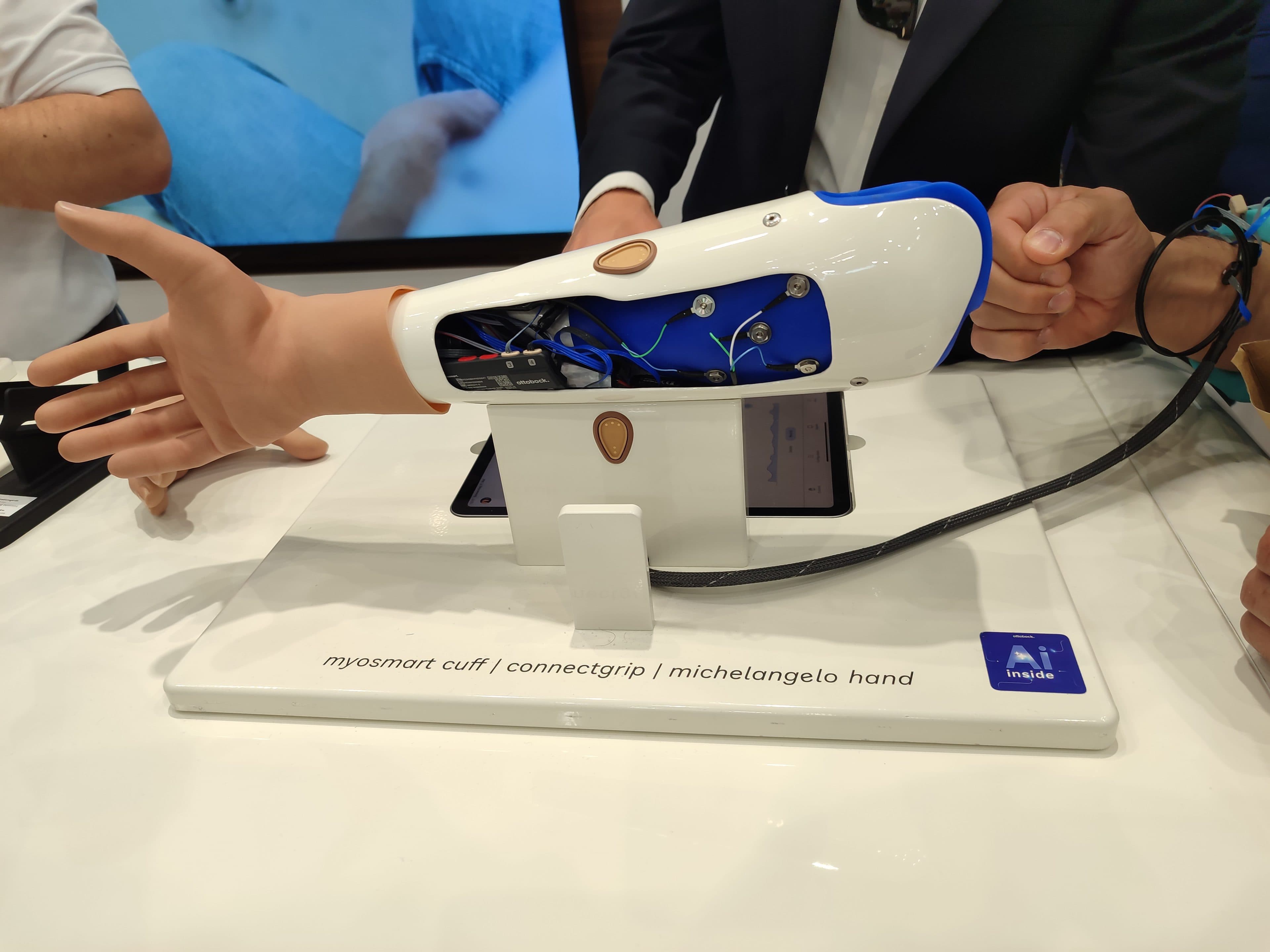 Eine Armprothese mit der Bezeichnung „Myosmart Cuff | ConnectGrip | Michelangelo Hand“ ist auf einem Ständer ausgestellt, sodass ihre inneren Mechanismen sichtbar sind, während Menschen sie betrachten und darauf zeigen.