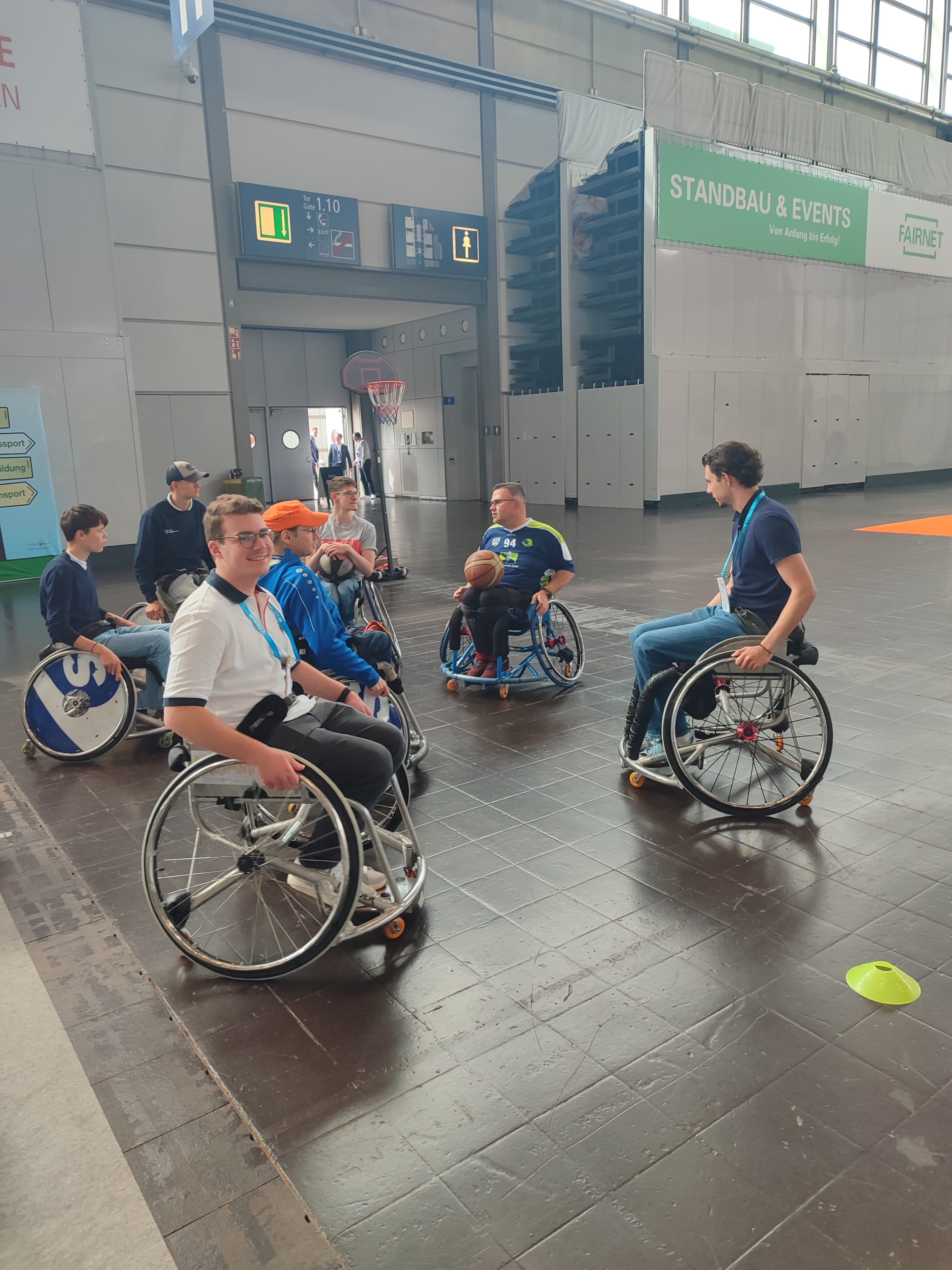 Eine Gruppe von Menschen im Rollstuhl spielt in einer Halle auf einem Basketballfeld mit einem Korb; einige halten Basketbälle in den Händen, während andere zuschauen oder mitspielen, und im Hintergrund sind große Veranstaltungsschilder zu sehen.