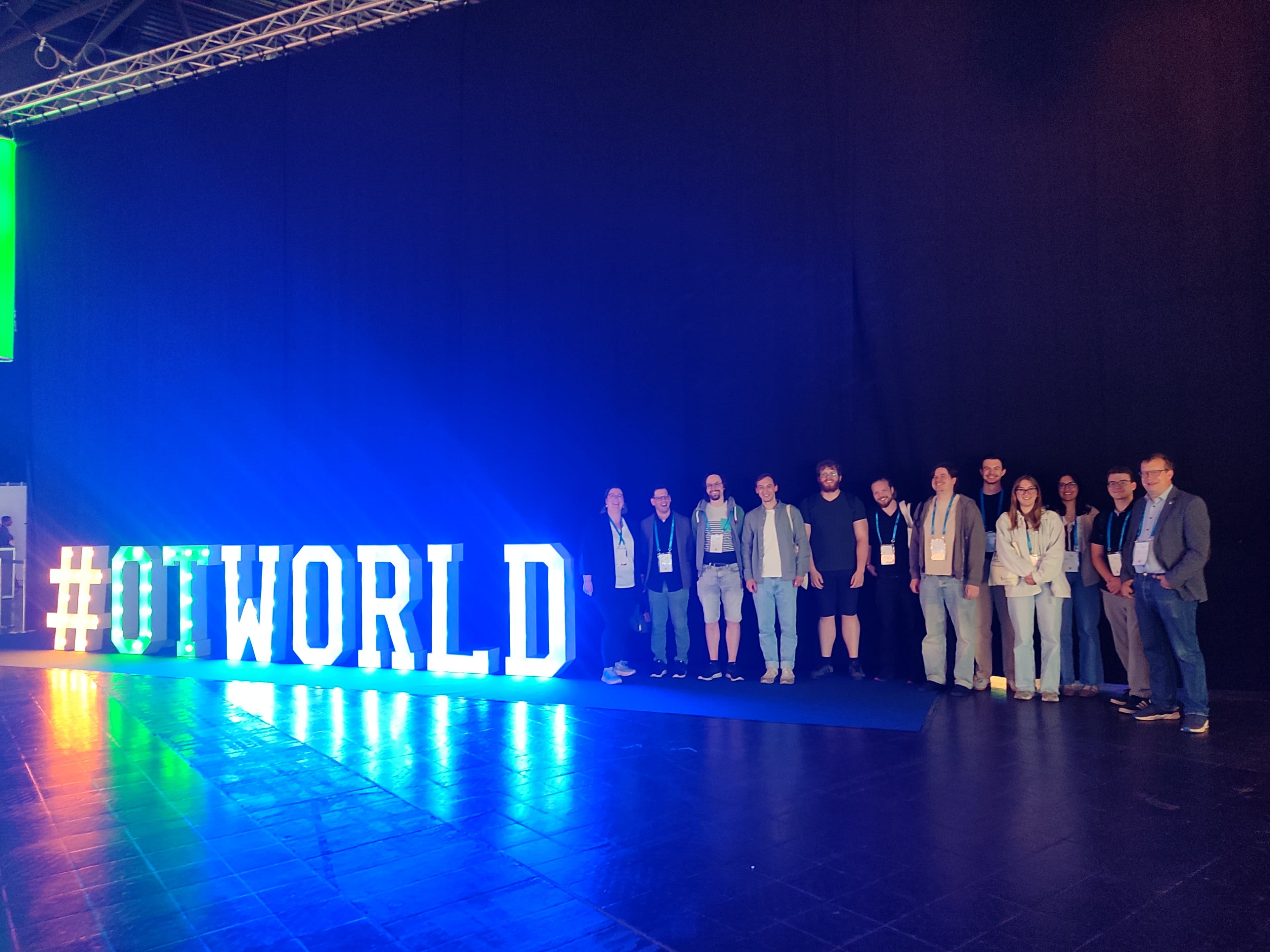 Bei einer Veranstaltung in einem Saal steht eine Gruppe von Menschen vor einem großen, beleuchteten #OTWORLD-Schild vor einem dunklen Hintergrund.