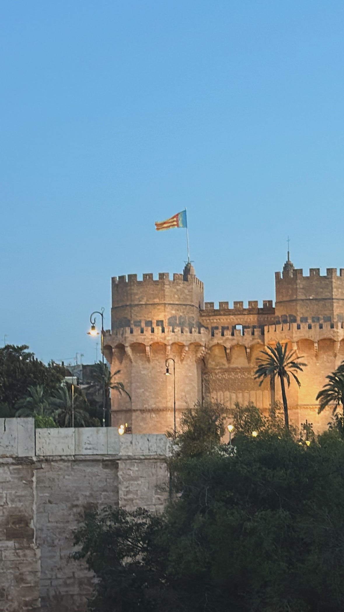 Eine historische Steinfestung mit Zinnentürmen und einer darauf wehenden Flagge, umgeben von Palmen, leuchtet in der Abenddämmerung vor einem strahlend blauen Himmel.