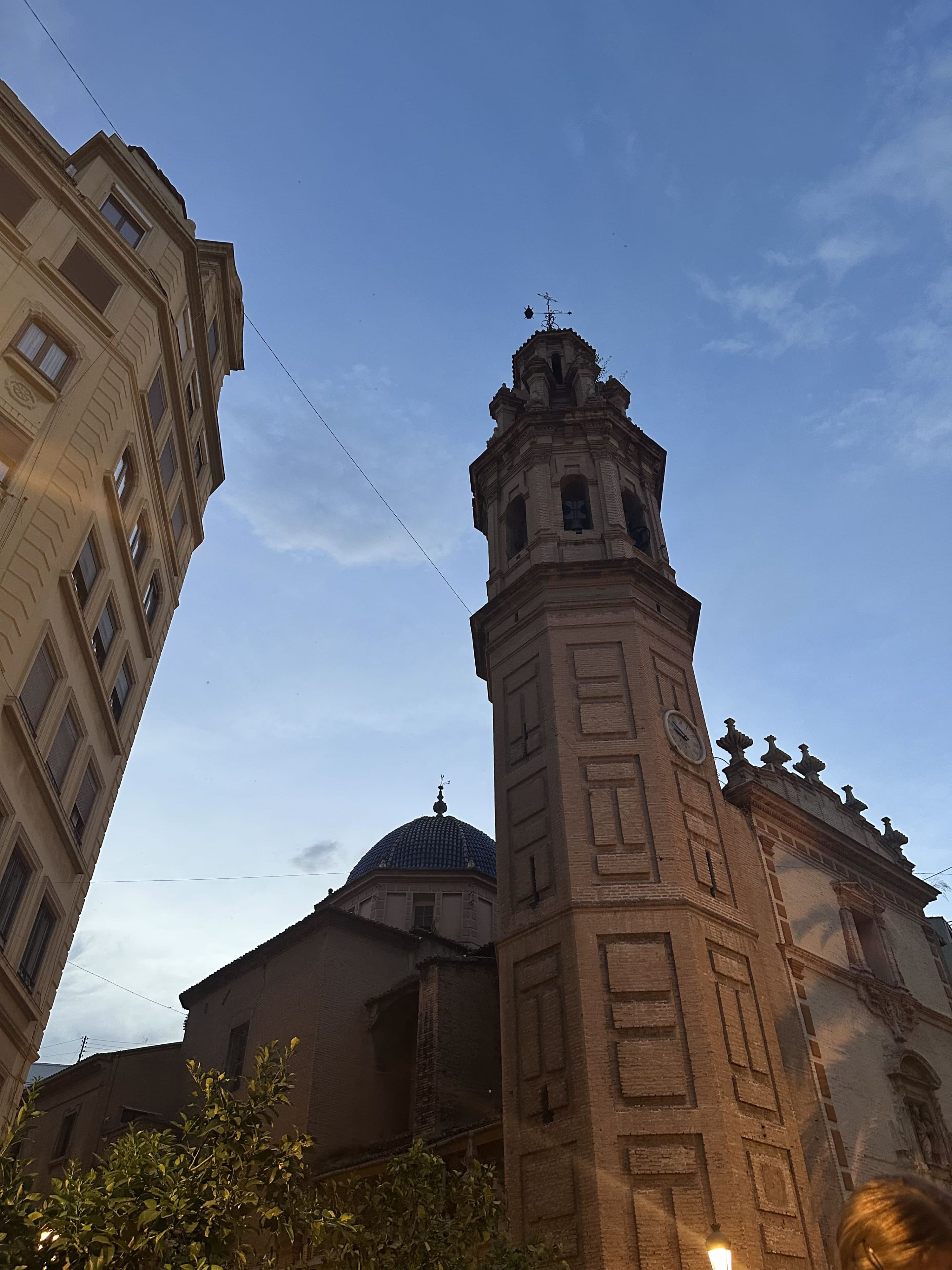Ein hoher, kunstvoll verzierter Glockenturm aus Backstein mit einer Uhr ragt vor einem blauen Abendhimmel empor, umgeben von historischen Gebäuden und einer Kuppelkirche. Am unteren Bildrand ist etwas Grün zu sehen.