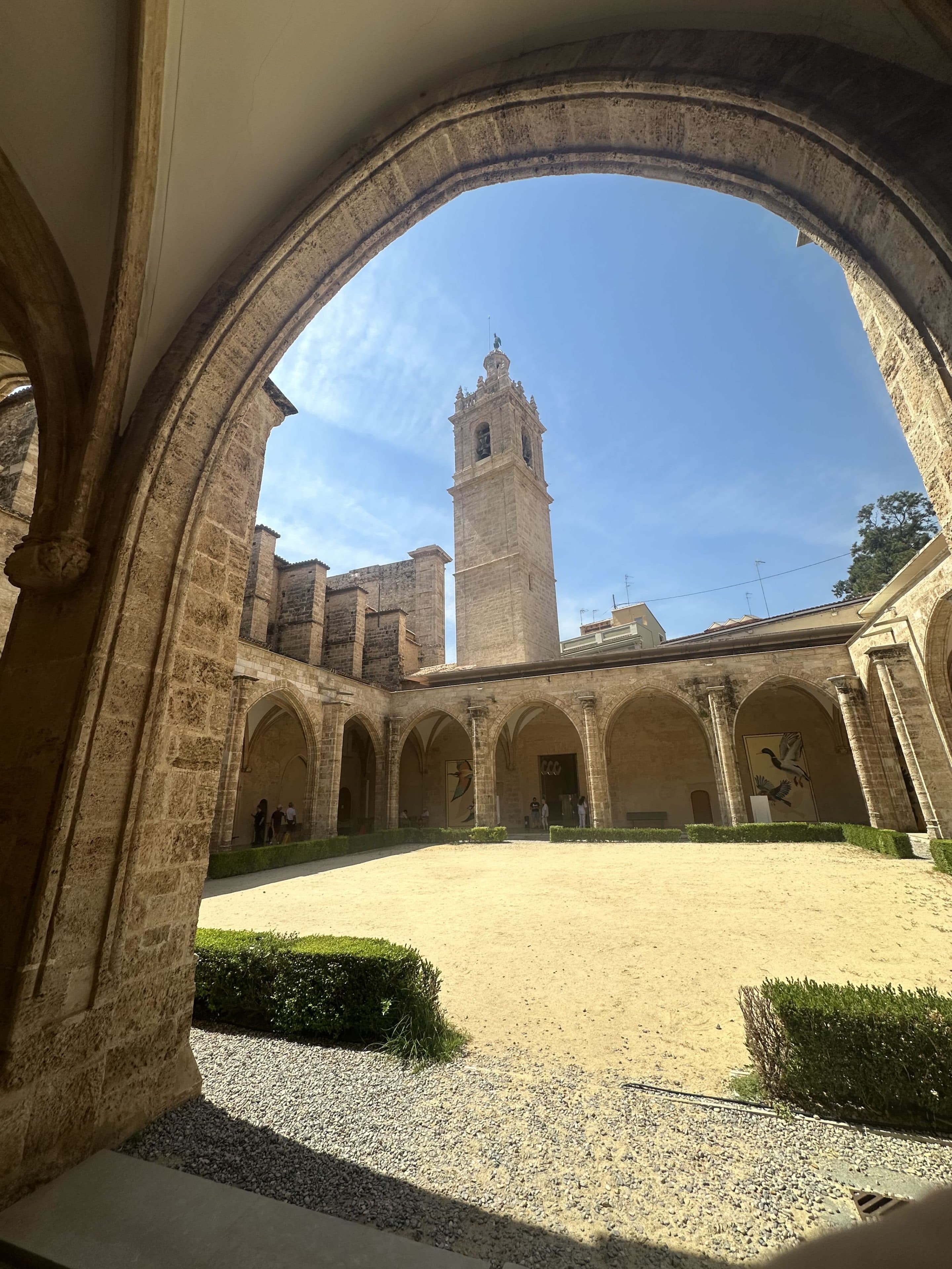 Ein Steinbogen umrahmt einen sonnigen Innenhof mit gepflegten Hecken und sandigem Boden, der von einem Säulenkreuzgang umgeben ist. Im Hintergrund ragt unter blauem Himmel ein hoher Glockenturm empor.