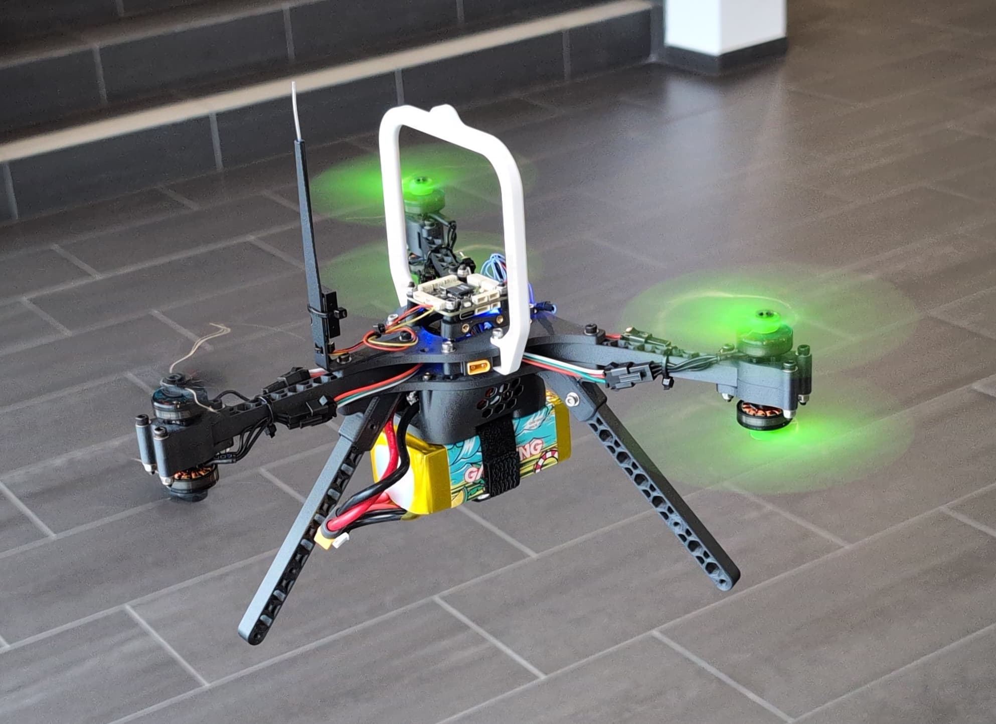 Eine Quadcopter-Drohne mit grünen LED-Leuchten an den Propellern schwebt in einem Innenraum über einem Fliesenboden. Die Drohne verfügt über freiliegende Kabel, einen Akku und einen weißen Griff an der Oberseite.