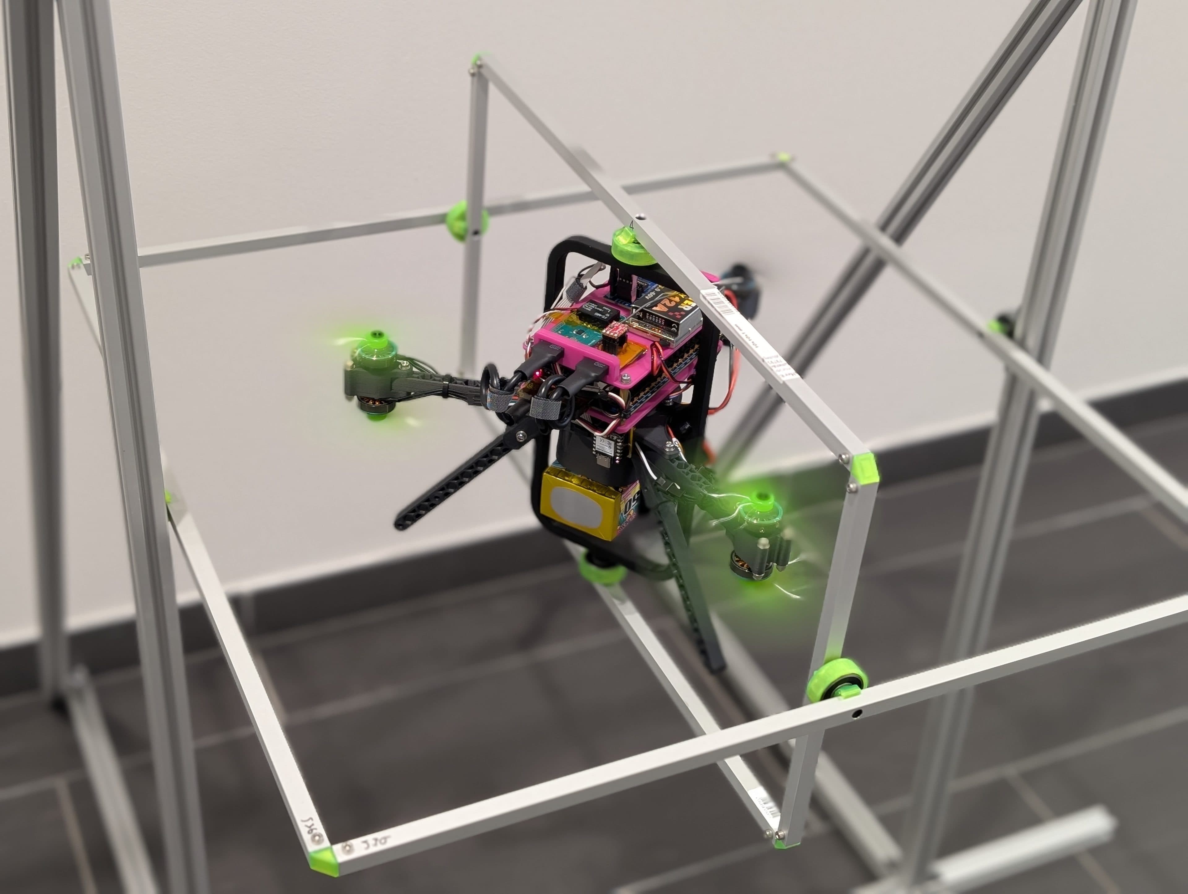 Eine Quadcopter-Drohne mit grün leuchtenden Propellern und verschiedenen elektronischen Bauteilen fliegt in einem Innenraum innerhalb einer Metallrahmenkonstruktion über einem dunklen Fliesenboden.