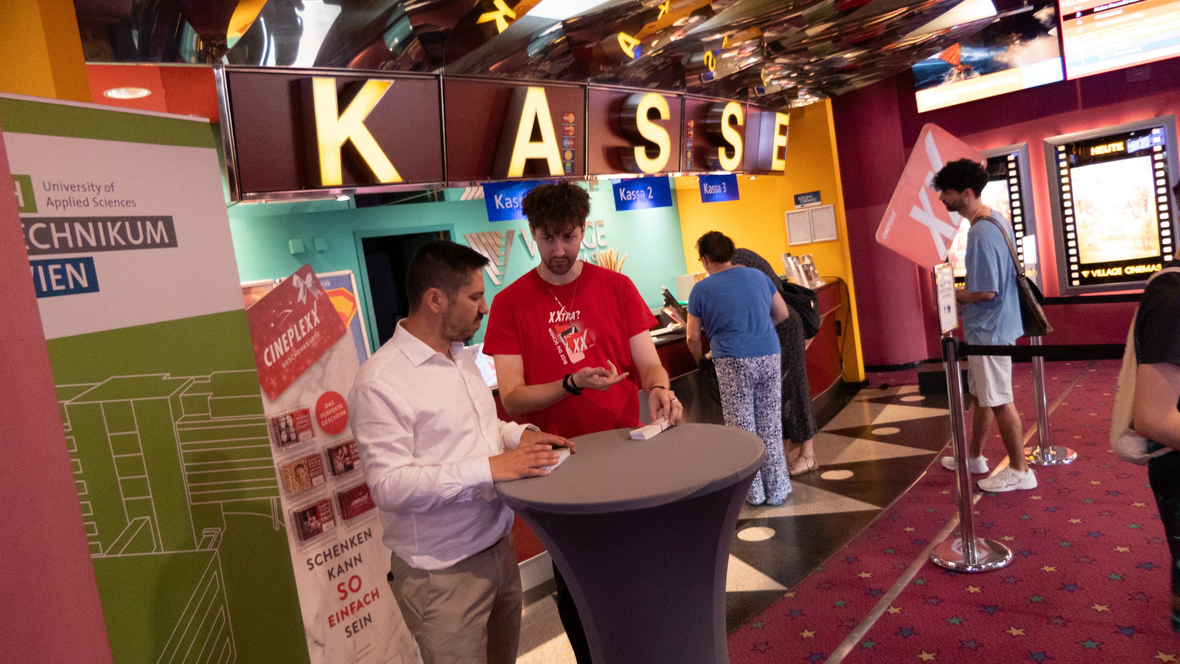 Zwei Männer stehen an einem runden Tisch und führen in der Nähe des Ticketschalters („KASSE“) eines Kinos einen Kartentrick vor. Andere Menschen warten in der Schlange, und bunte Plakate und Banner schmücken den Bereich.