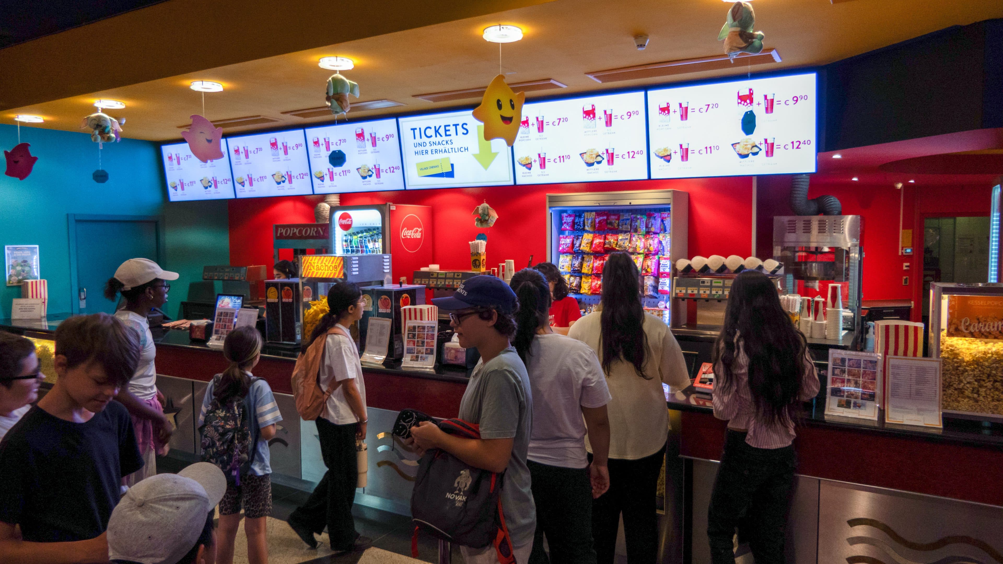 Die Leute stehen an einem farbenfrohen Imbissstand in einem Kino Schlange, über dem leuchtende Menüs mit Snacks und Getränken zu sehen sind. Auf der Theke und hinter den Mitarbeitern sind verschiedene Sorten Popcorn, Süßigkeiten und Limonaden zu sehen.
