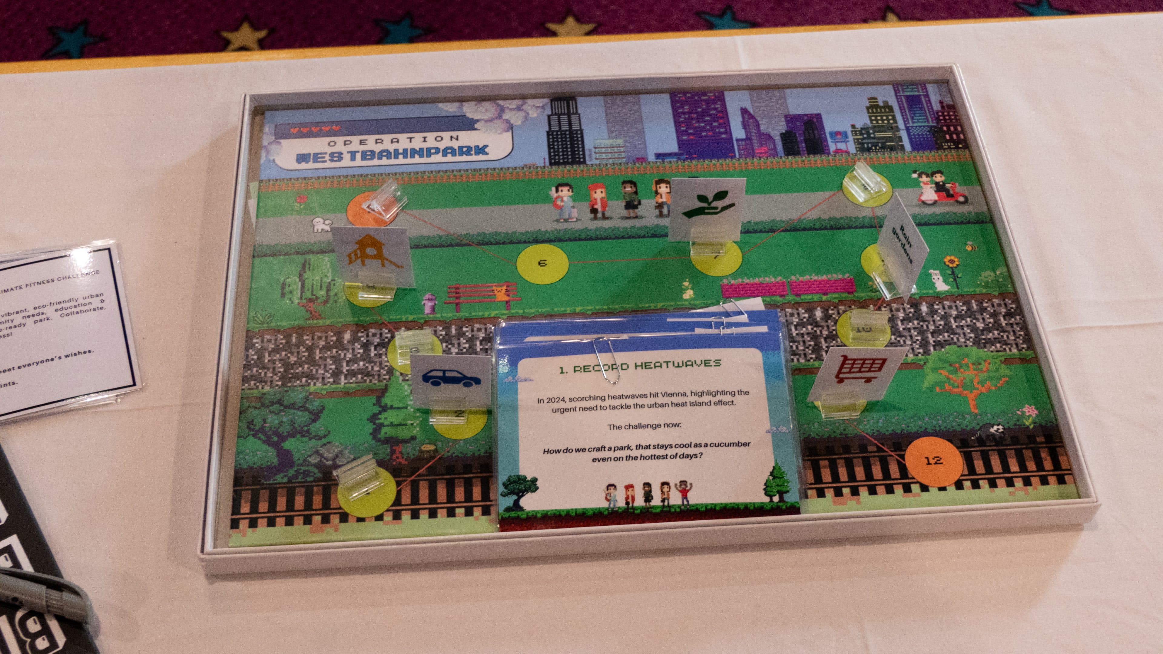 Auf einem Tisch steht ein farbenfrohes Brettspiel mit dem Titel „Operation Westbahnpark“, das auf einem Spielbrett mit Parkmotiv illustrierte Figuren, Symbole, Textkarten und Zahlenmarken zeigt.