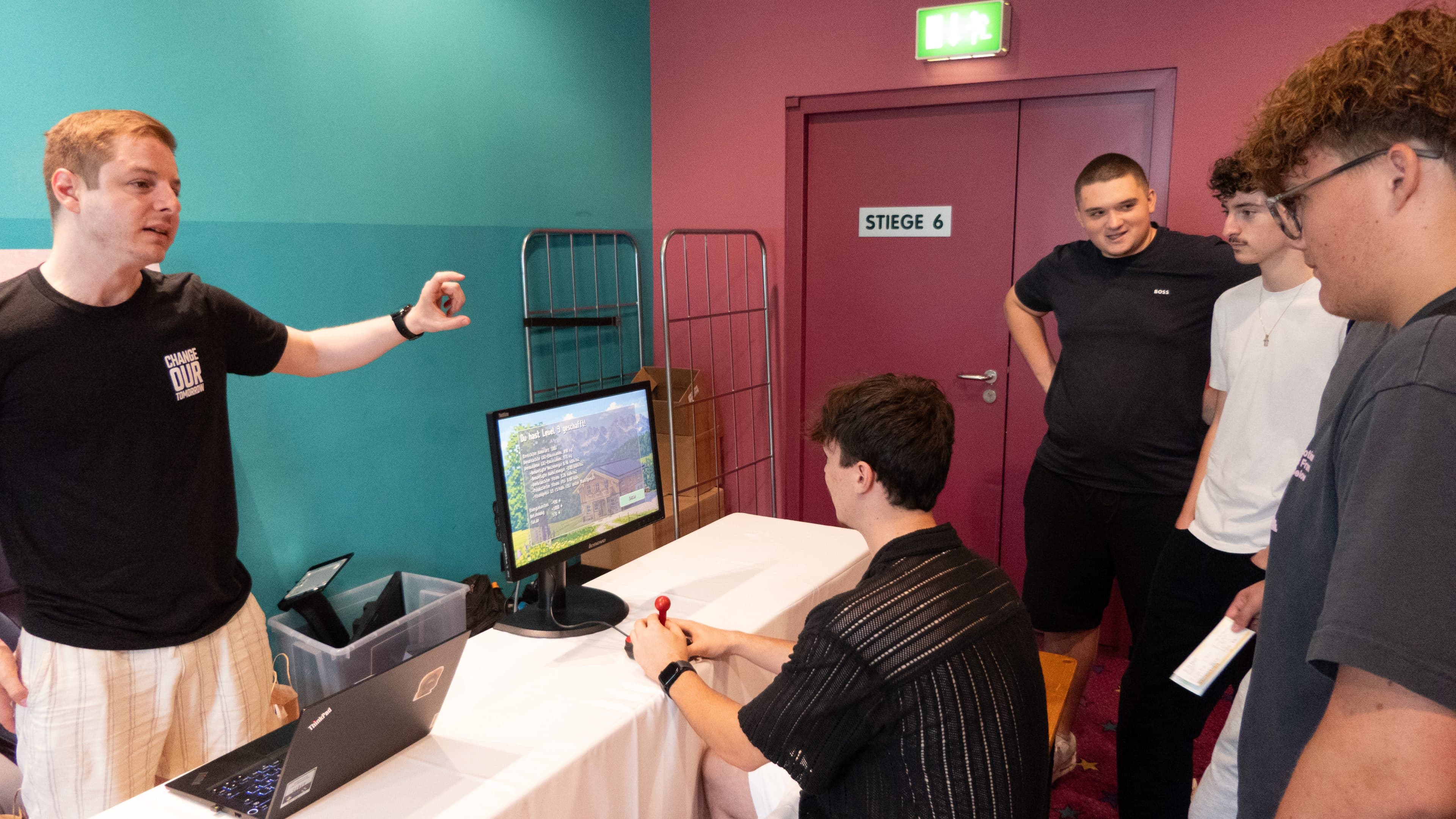 Eine Gruppe junger Männer versammelt sich um einen Tisch, an dem einer ein Spiel am Computer spielt, während ein anderer gestikuliert und etwas erklärt. Vier weitere schauen aufmerksam zu – in einem farbenfrohen Raum mit türkis- und rotgestrichenen Wänden.