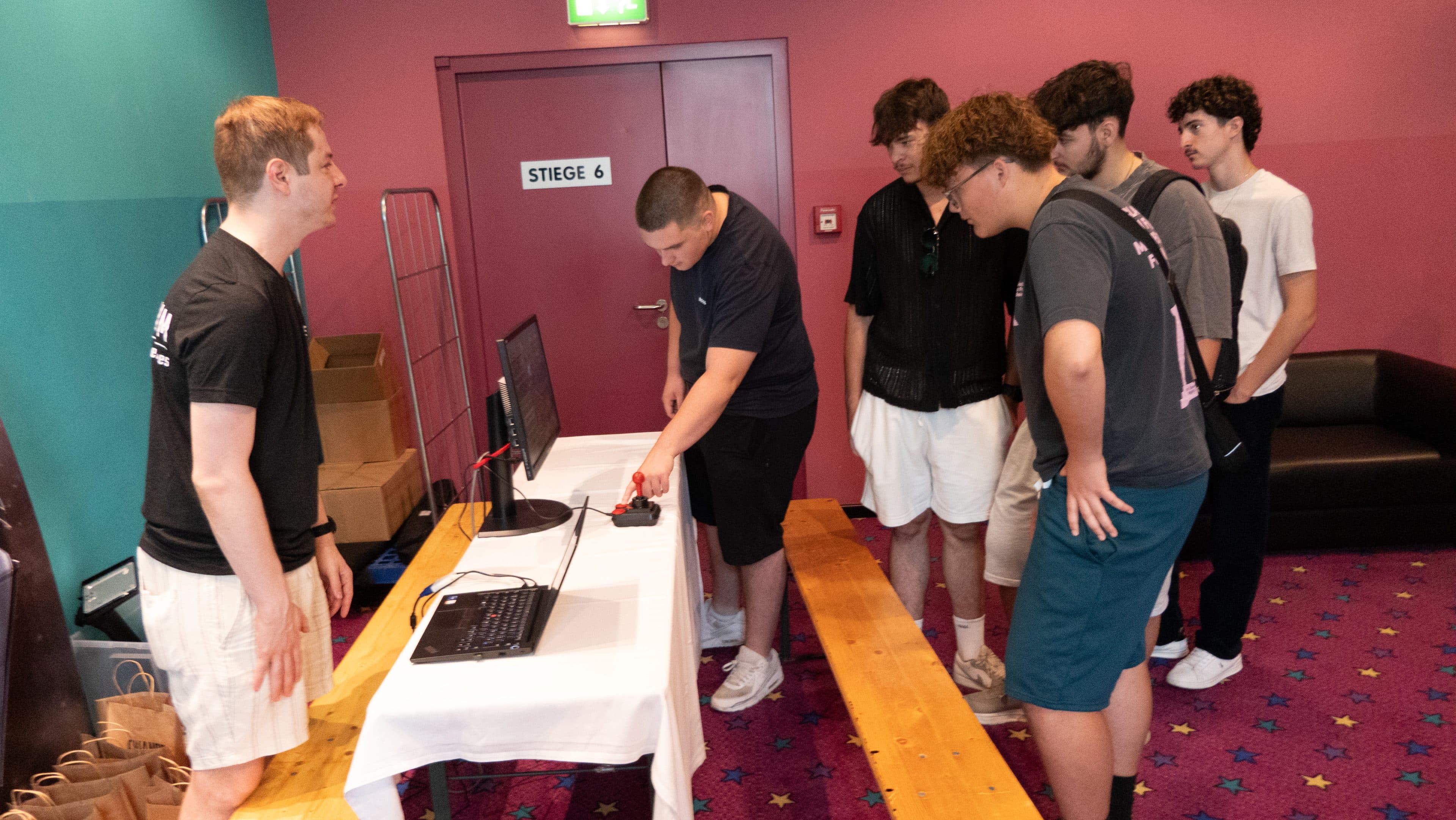 Sechs junge Männer stehen um einen Tisch mit einem Monitor und einem Joystick herum, während einer von ihnen mit dem Gerät interagiert und ein anderer, der auf der gegenüberliegenden Seite steht, zusieht und lächelt. Der Schauplatz scheint ein Veranstaltungsraum zu sein.
