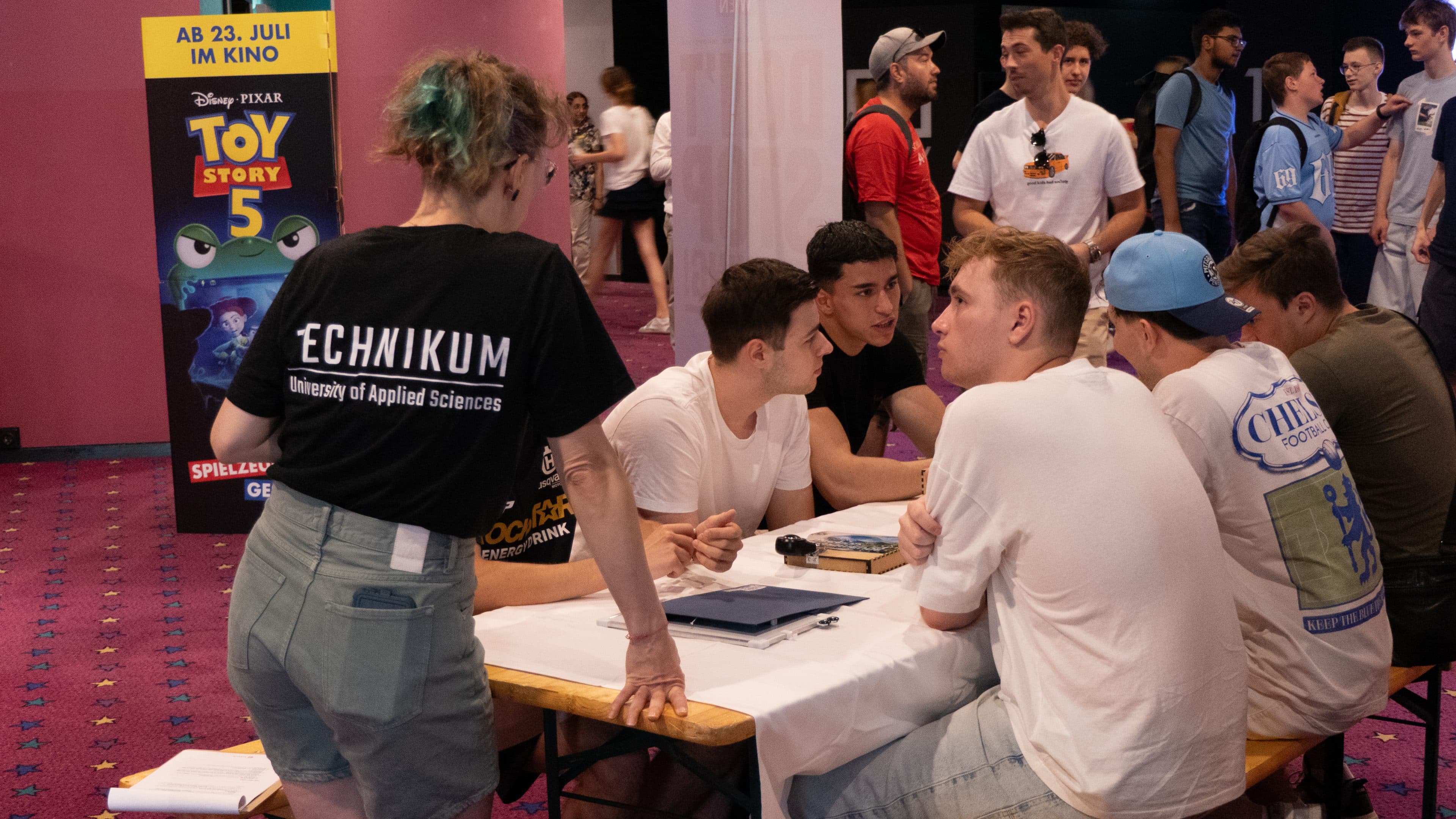 Eine Gruppe junger Männer sitzt um einen Tisch herum und unterhält sich mit einer Frau, die ein schwarzes T-Shirt der Fachhochschule Technikum trägt. Im Hintergrund sind ein Filmplakat zu „Toy Story 5“ und einige Personen zu sehen.