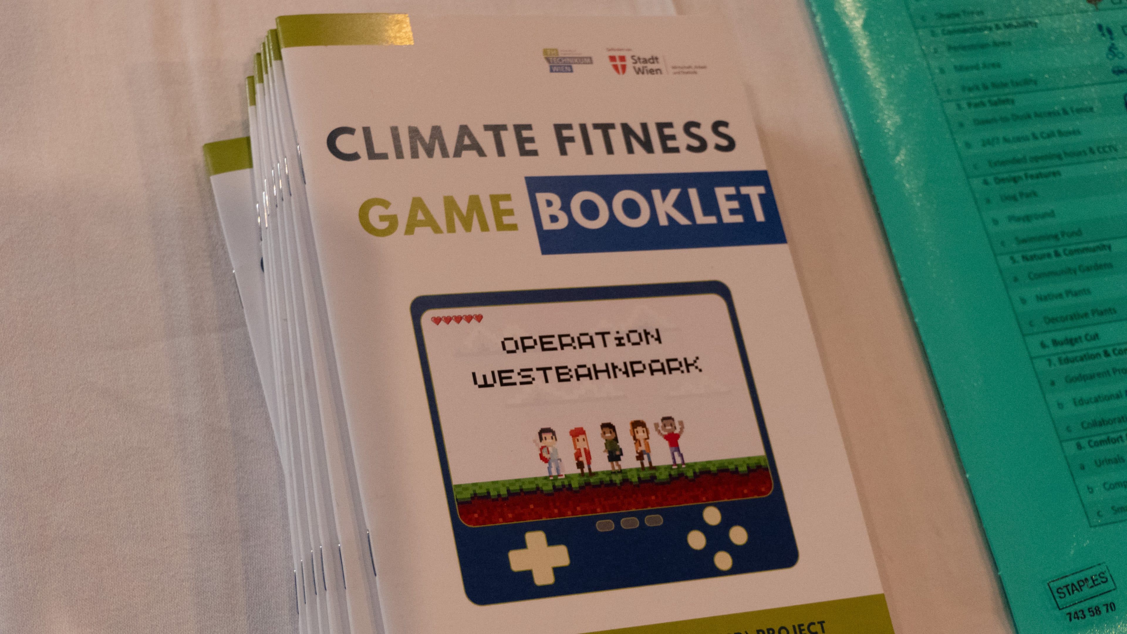 Ein Stapel Broschüren zum „Climate Fitness Game Booklet“ mit einem Spielbildschirm im Pixel-Art-Stil, betitelt „Operation Westbahnpark“, sowie Comicfiguren und der Abbildung eines Spielcontrollers auf dem Cover.