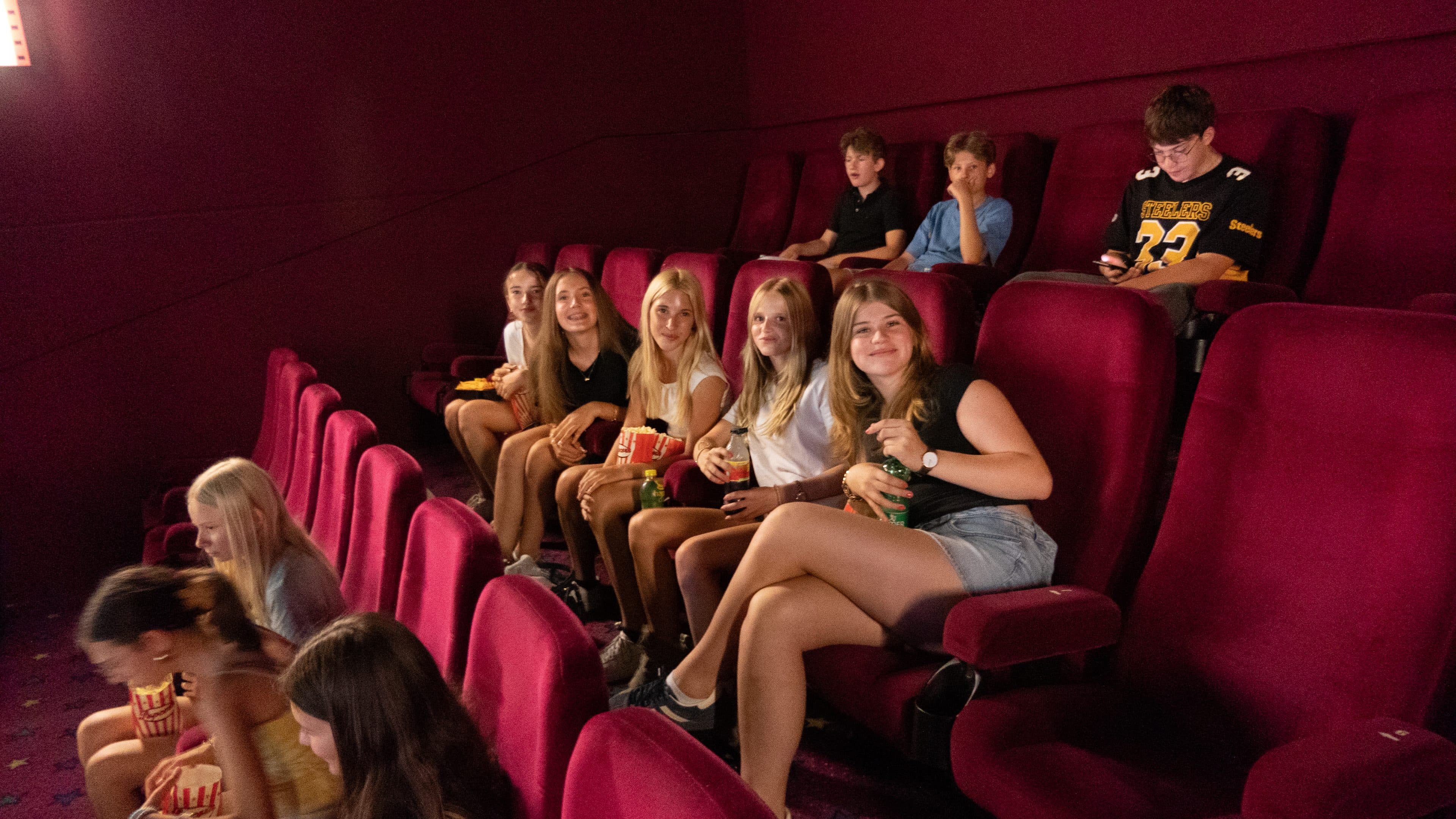 Eine Gruppe von Teenagern sitzt auf roten Kinosesseln, lächelt und hält Popcorn in den Händen, während einige andere verstreut in der Nähe sitzen. Der Kinosaal ist fast leer, und die Beleuchtung ist warm.