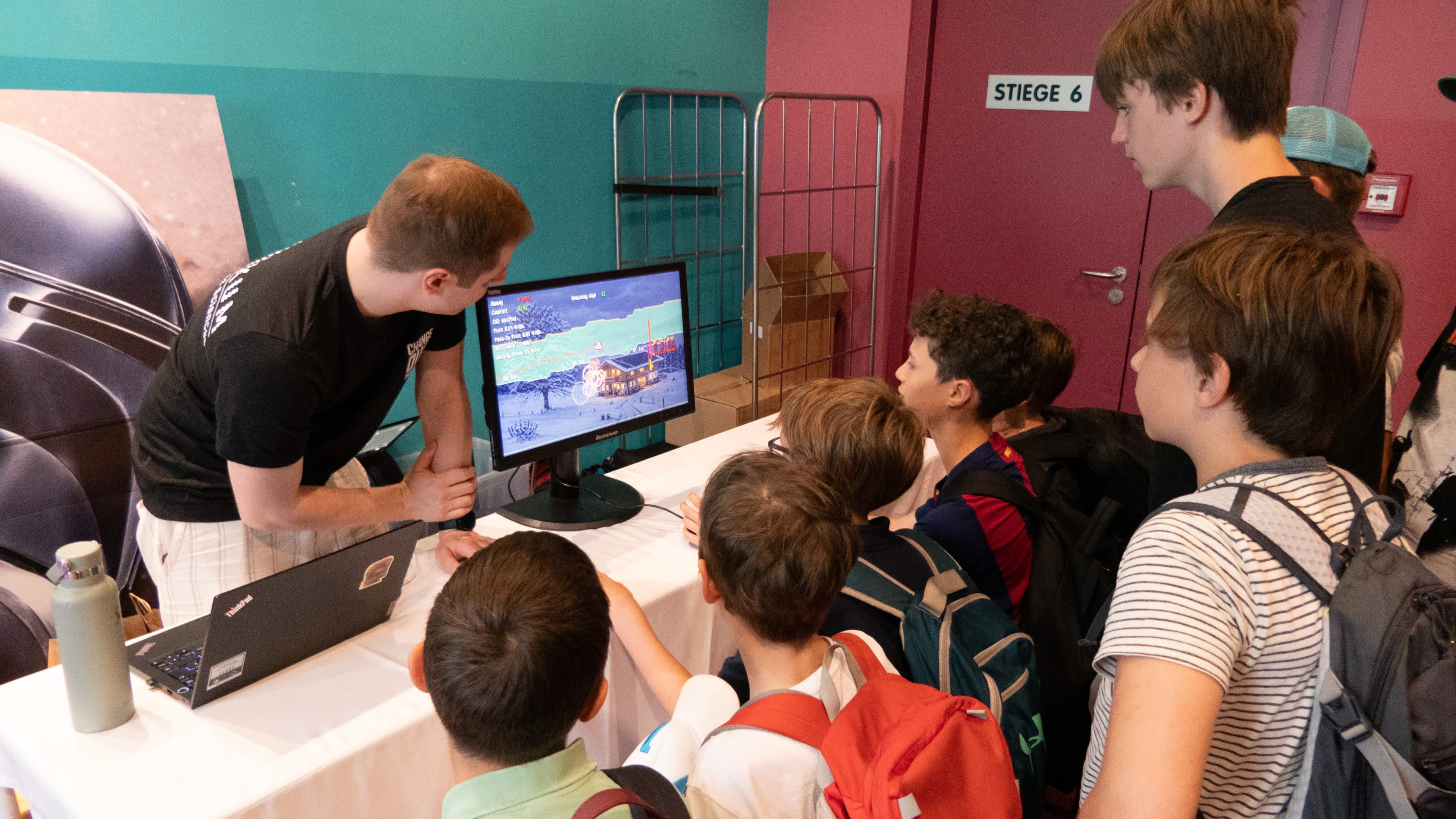 Ein Mann führt einer Gruppe von Kindern und Jugendlichen, die sich bei einer Veranstaltung in einem Saal um einen Tisch versammelt haben, ein Videospiel auf einem Monitor vor. Die Gruppe hört aufmerksam zu, und bei einigen Kindern sind Rucksäcke zu sehen.