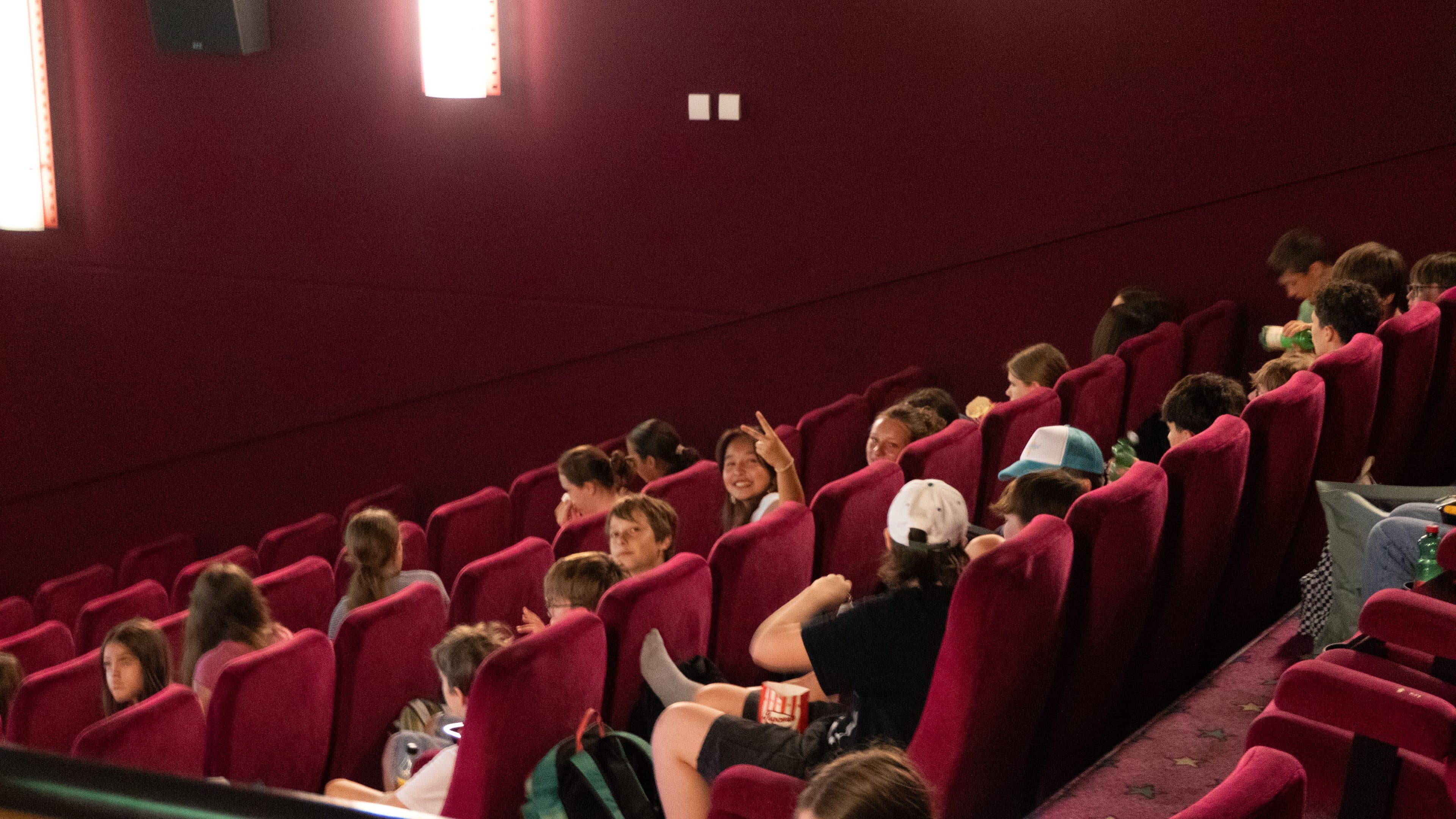 Eine Gruppe von Kindern sitzt in Reihen roter Sitze in einem schummrig beleuchteten Kino; einige unterhalten sich und lächeln, während andere nach vorne schauen oder Snacks essen.