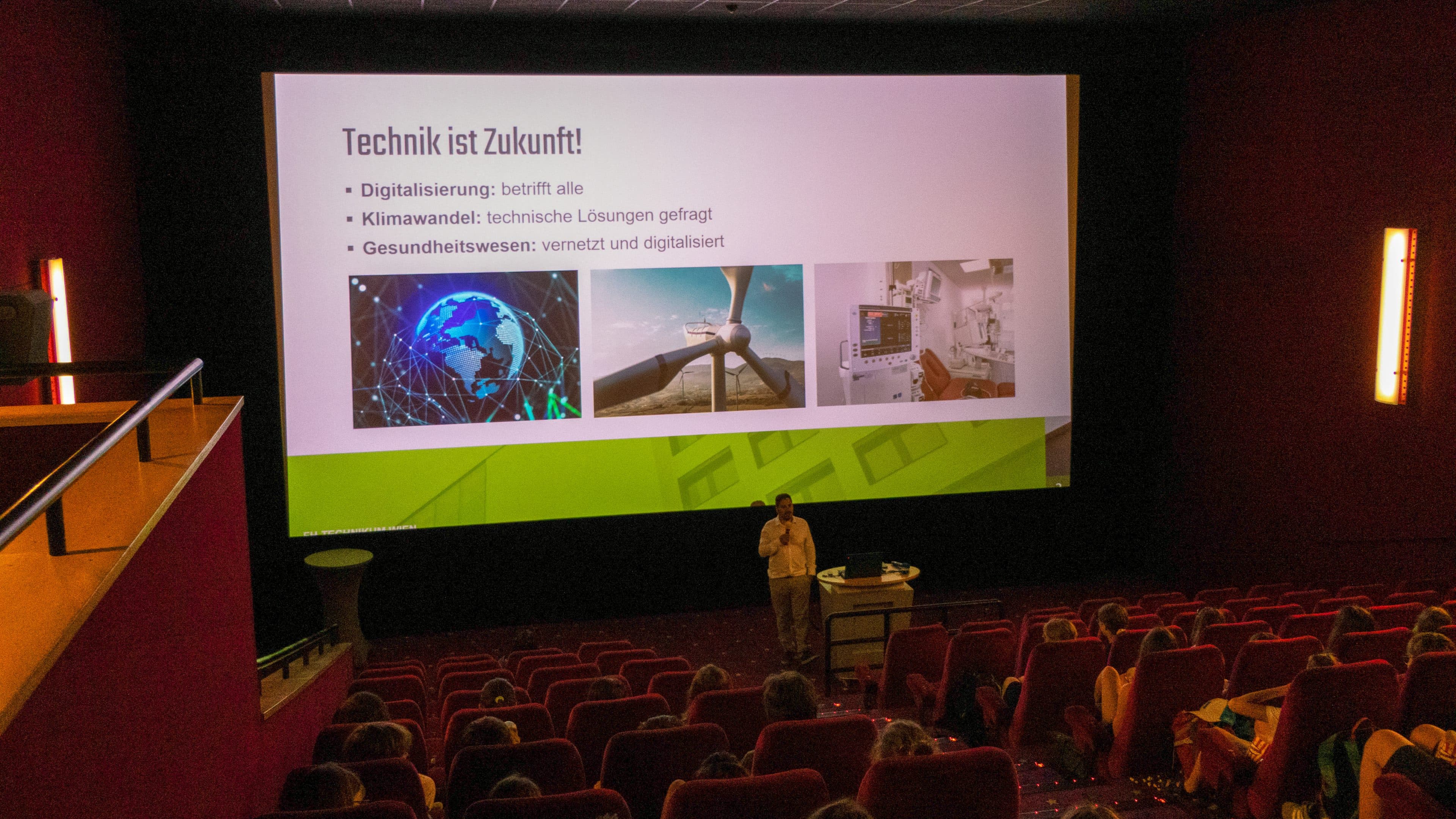 Ein Redner hält einen Vortrag in einem abgedunkelten Kinosaal. Auf der Leinwand ist eine deutsche Folie mit der Überschrift „Technik ist Zukunft!“ zu sehen, die Bilder zu den Themen Technik, Klimawandel und Gesundheitswesen zeigt, während das Publikum von roten Sitzen aus zusieht.