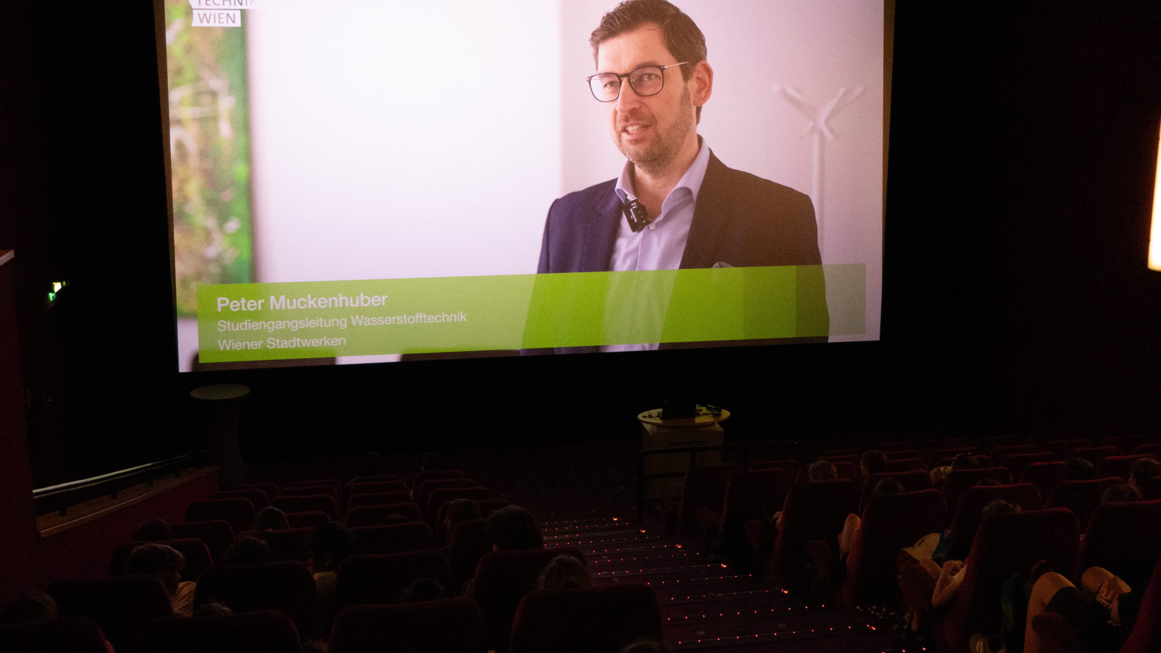 Ein Kinopublikum blickt auf eine große Leinwand, auf der ein Mann mit Brille und Anzug zu sehen ist, der als Peter Muckenhuber vorgestellt wird und in einer Videopräsentation über Wasserstofftechnologie spricht.