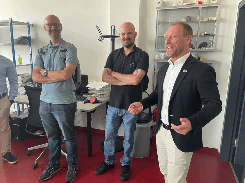 Drei Männer stehen in einem hellen Büroraum mit rotem Bodenbelag; einer von ihnen, der einen Blazer trägt, spricht und gestikuliert, während die beiden anderen, die Freizeithemden und Jeans tragen, zuhören und lächeln. Im Hintergrund sind Schreibtische und Regale mit verschiedenen Gegenständen zu sehen.