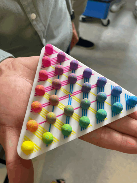 Eine Person hält ein dreieckiges Spielbrett in der Hand, auf dem bunte, runde Spielsteine und sich kreuzende Neonlinien ein geometrisches Muster bilden. Die Spielsteine sind nach Farben in Reihen angeordnet; das Brett scheint im 3D-Druckverfahren hergestellt worden zu sein.