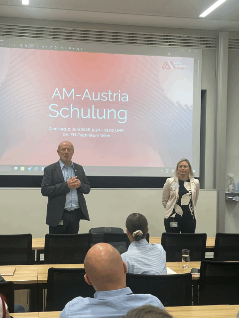 Ein Mann und eine Frau stehen vorne im Klassenzimmer und halten einen Vortrag über „AM-Austria Schulung“. Die Teilnehmer sitzen und hören zu. Auf einer großen Leinwand werden Veranstaltungsdetails auf Deutsch angezeigt.