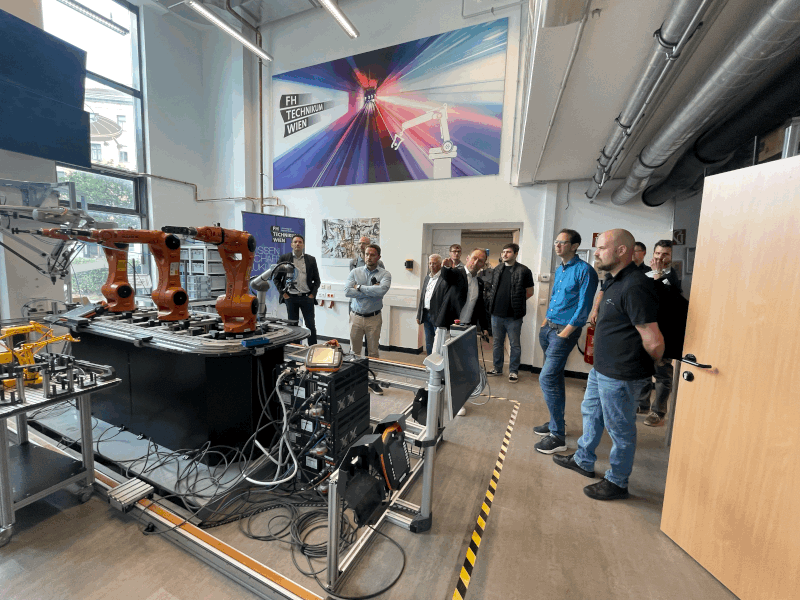 Eine Gruppe von Menschen beobachtet Roboterarme bei der Arbeit in einem modernen Industrielabor mit großen Fenstern, Maschinen und einem bunten Technologieplakat an der Wand.