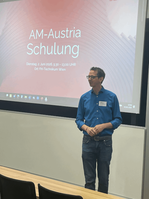 Ein Mann in einem blauen Hemd steht vor einer Präsentationswand mit der Aufschrift „AM-Austria Schulung“ und Veranstaltungsdetails auf Deutsch an der FH-Technikum Wien. Er scheint sich an das Publikum zu wenden.