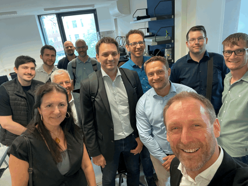Eine Gruppe von dreizehn Erwachsenen, überwiegend Männer, die lächelnd gemeinsam in einem Büro oder Labor posieren, im Hintergrund sind Computer und Geräte zu sehen.