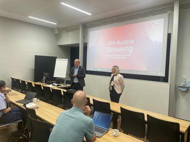 Zwei Personen stehen vorne im Klassenzimmer und halten einen Vortrag mit dem Titel „AM-Austria Schulung“. Die Teilnehmer sitzen an Tischen mit Laptops und Kaffeetassen und blicken auf die Referenten und eine Leinwand.