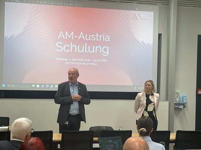 Ein Mann und eine Frau stehen vorne im Klassenzimmer und halten einen Vortrag mit dem Titel „AM-Austria Schulung“, hinter ihnen wird eine Folie projiziert; mehrere Personen sitzen ihnen gegenüber.