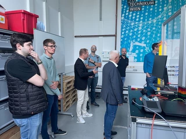 Eine Gruppe von sieben Männern steht in einer Technikwerkstatt und verfolgt eine Vorführung an einem Arbeitsplatz mit Bildschirmen und Geräten. Im Raum befinden sich Regale, ein Whiteboard und ein großes blaues Wandplakat mit Robotermotiven.