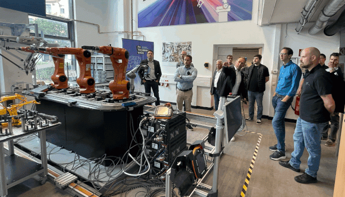 Eine Gruppe von Menschen steht in einem modernen Labor und betrachtet Roboterarme und Maschinen auf Werkbänken. Der Raum ist mit Industrieanlagen, freiliegenden Rohrleitungen und einem großen Poster mit Technikmotiven an der Wand ausgestattet.