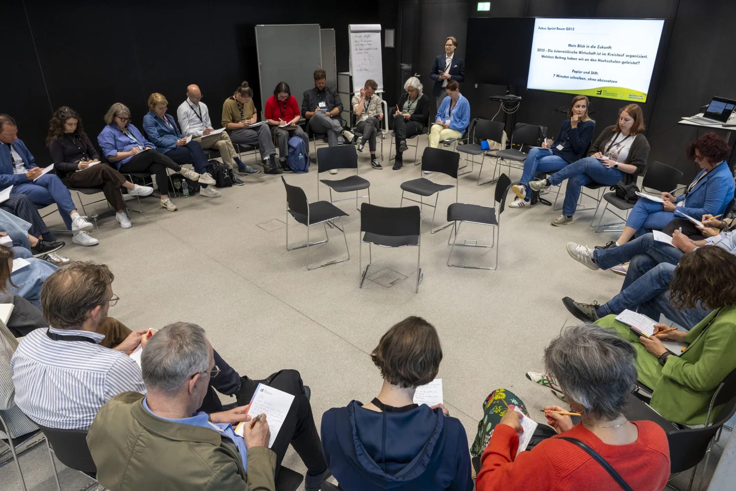 Eine Gruppe von Menschen sitzt in einem modernen Raum im Kreis, macht sich Notizen und hört einem Redner zu, der neben einer Leinwand steht, auf der eine Präsentation zu sehen ist. In der Mitte des Kreises stehen mehrere leere Stühle.