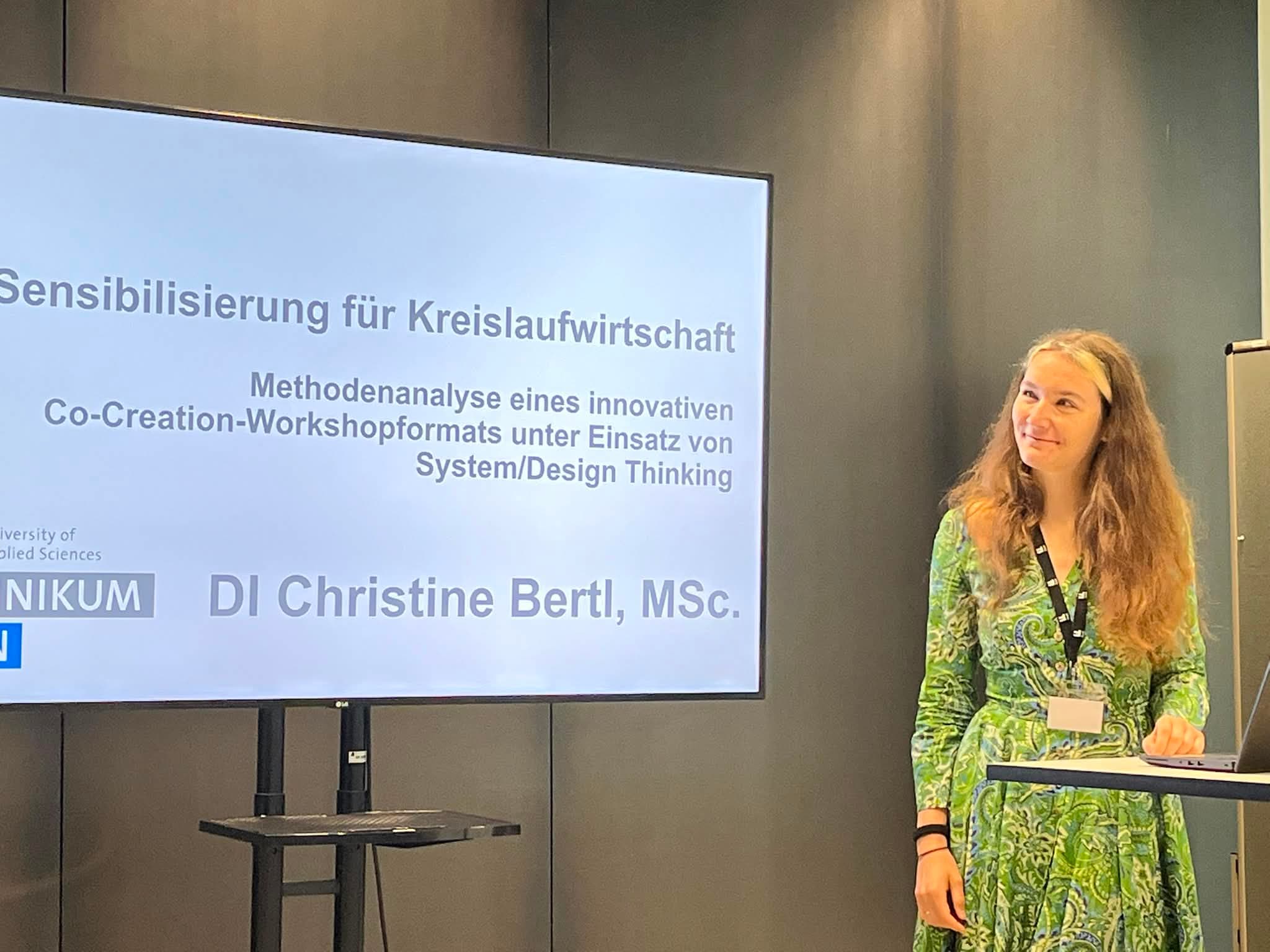 Eine Frau in einem grünen Kleid steht neben einer Leinwand, auf der eine deutsche Präsentation mit dem Titel „Sensibilisierung für Kreislaufwirtschaft“ von DI Christine Bertl, MSc, zu sehen ist. Sie lächelt und hält einen Klicker in der Hand.