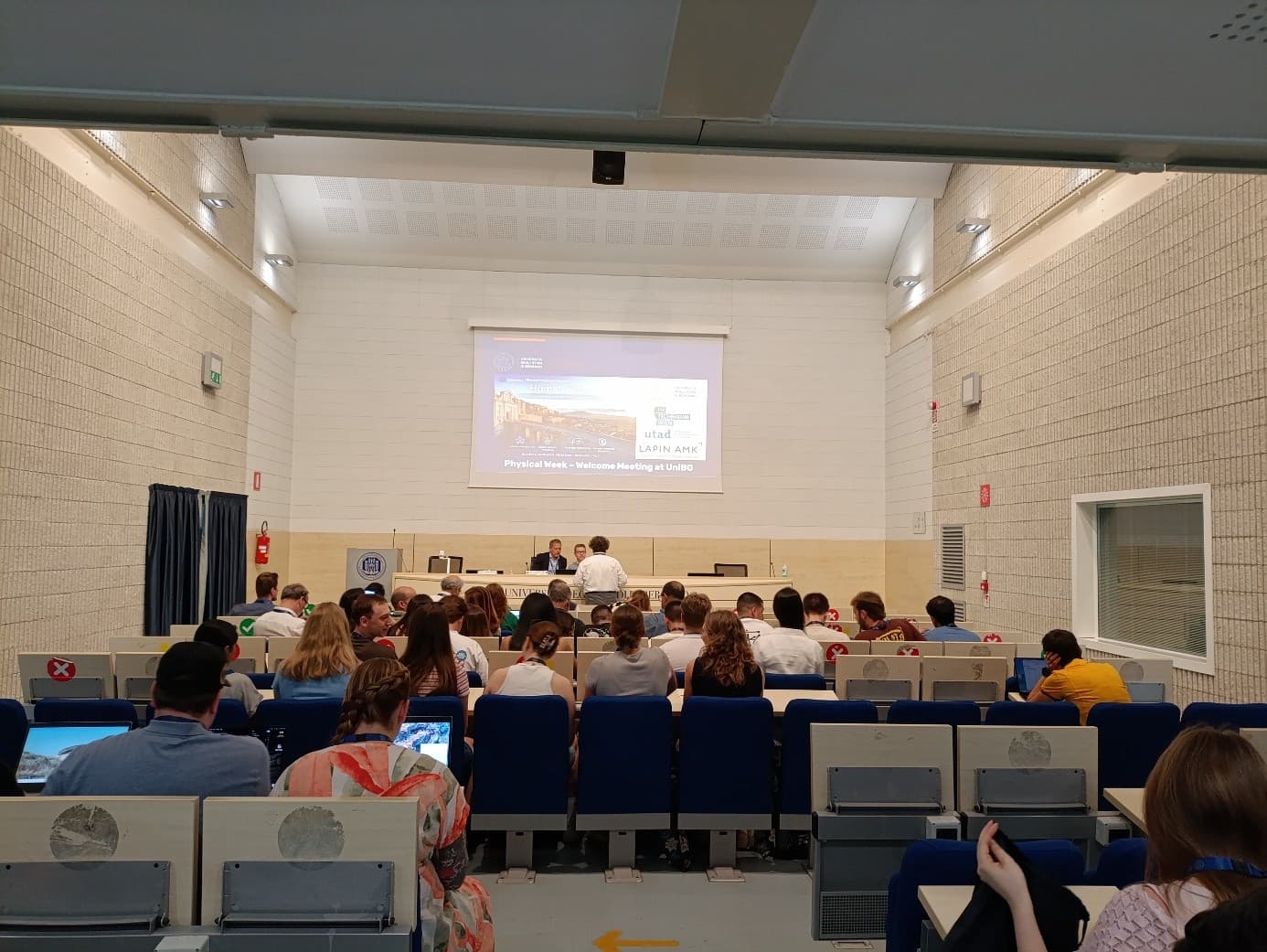 Ein Klassenzimmer, in dem die Schüler an ihren Schreibtischen sitzen und auf eine Projektionswand blicken, auf der eine Präsentation gezeigt wird. Der Raum hat beigefarbene Wände und blaue Stühle; vorne steht ein Dozent, der zu den Zuhörern spricht.