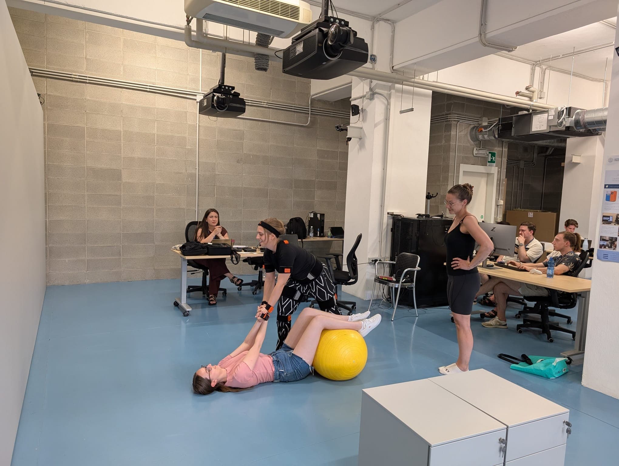 Eine Frau, die auf einem gelben Gymnastikball liegt, wird von einer anderen Frau unterstützt, während andere zuschauen oder an Schreibtischen in einem modernen Klassenzimmer mit blauem Boden und freiliegenden Rohrleitungen arbeiten.
