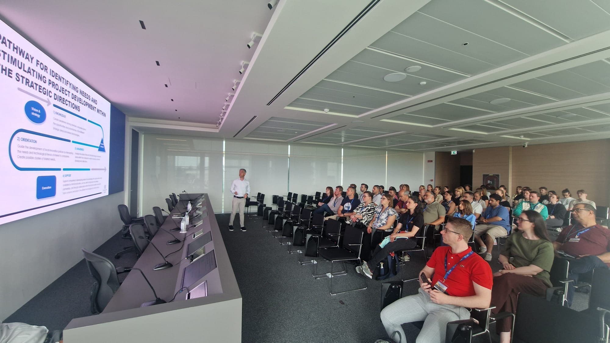 Eine Person steht vorne in einem modernen Konferenzraum und hält vor einem Publikum einen Vortrag. Auf einer großen Leinwand ist ein Flussdiagramm zu sehen, während die Teilnehmer in Reihen sitzen und aufmerksam zuschauen und zuhören.