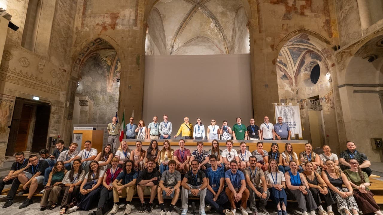 Eine große Gruppe von Menschen posiert für ein Foto in einem historischen, kunstvoll bemalten Saal mit hohen Gewölbedecken und Steinwänden. Die Gruppe sitzt und steht auf gestuften Bänken und lächelt in die Kamera.